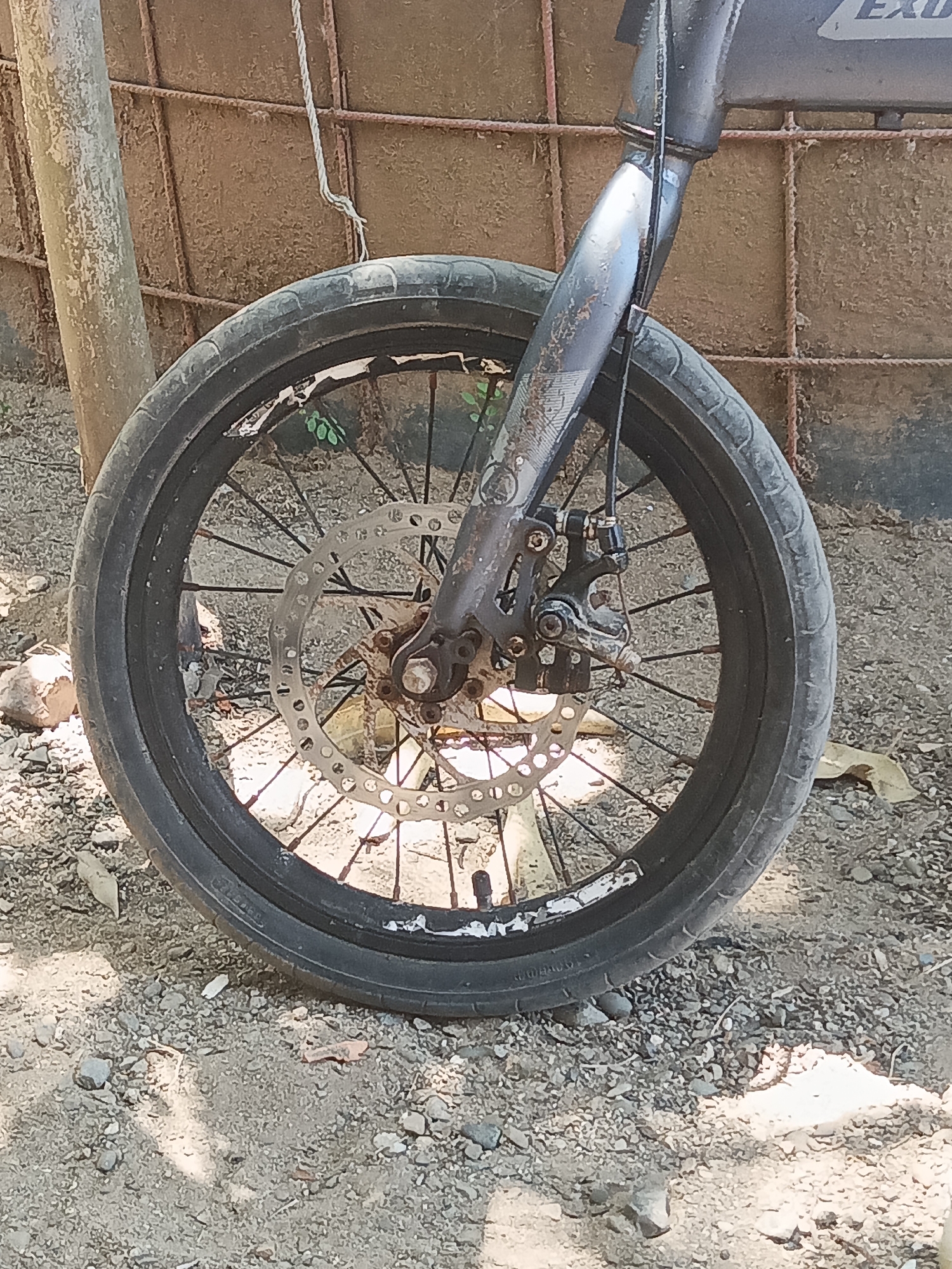 The image shows the front wheel and fork of a bicycle. The bicycle appears to be parked outdoors, with a rough, gravelly ground surface. In the background, there is a rusty metal fence and a wooden post. There is no indication of the city of Pitue, Indonesia, in the image.