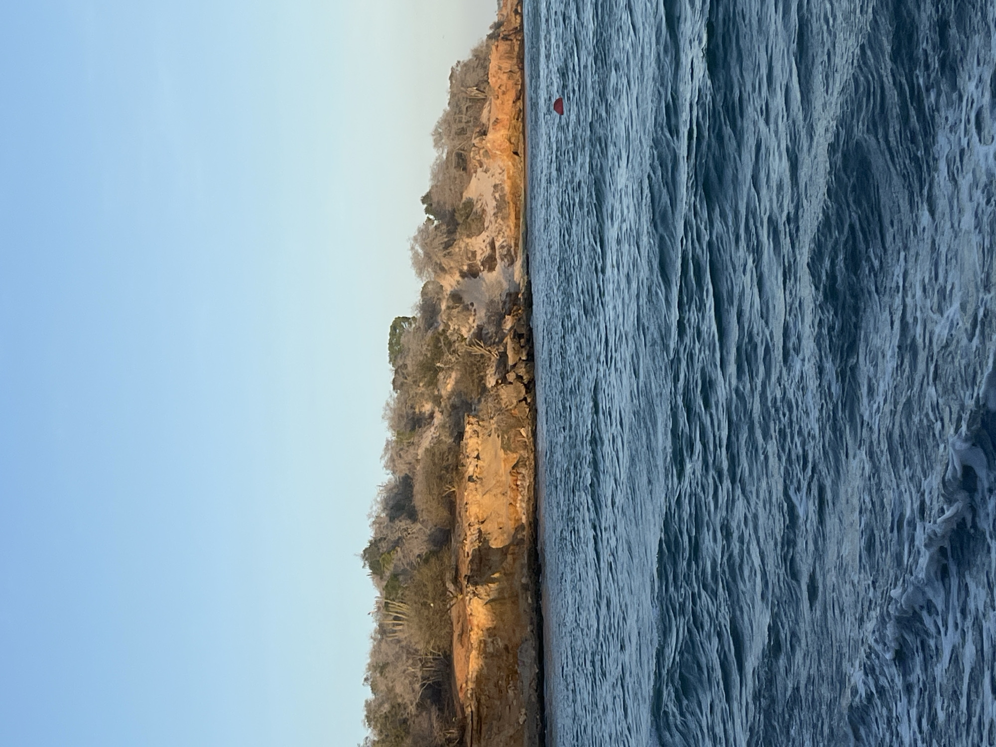 A photograph shows a landscape, oriented vertically with the sky on the left. The sky is light blue, transitioning to pale yellow towards the top. Adjacent to the sky is a cliff face with vegetation atop. The cliff shows visible erosion and layering.
To the right of the cliff is the ocean. The surface is ruffled with wave action. A red buoy is visible in the water, slightly offshore from the cliff.
The location is suggested to be El Yaque, Venezuela, based on the request.