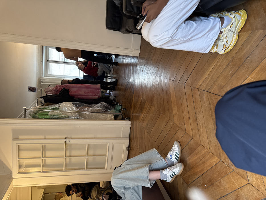 The image depicts an interior scene, characteristic of a Parisian apartment, likely a Haussmannian-style building in Paris, France, identifiable by its ornate chevron parquet flooring and tall white paneled doors.
In the immediate foreground, the lower extremities of three individuals are visible. To the left, a person wears light blue wide-leg jeans and white Adidas Samba sneakers with green stripes. To the right, a person in white wide-leg pants wears yellow chunky athletic sneakers and holds a silver-colored smartphone. The lower portion of a third person, clad in dark blue fabric, occupies the bottom center.
The midground reveals a wide doorway leading into an adjacent room. To the left of this opening, a white paneled door with multiple glass inserts is partially open. Across the doorway, a mobile metal clothing rack is laden with various garments, some encased in transparent plastic, prominently featuring a pink patterned dress. A person in dark attire stands near this rack.
Through the doorway, the background room contains several individuals and objects. A man with a bare torso, wearing dark pants, stands with his back partially to the viewer. To his left, a man in a red long-sleeved shirt is seated, engaged in interaction. A woman with blonde hair is also visible within this group. Further left, a man with dark curly hair and sunglasses, dressed in a dark jacket, is seated and viewing a smartphone. A photograph or print of a woman's face is taped to the wall above the clothing rack. A large window in the background room provides natural light. The arrangement of clothing racks and multiple individuals suggests a professional preparation or backstage environment, possibly related to a fashion fitting or photoshoot.