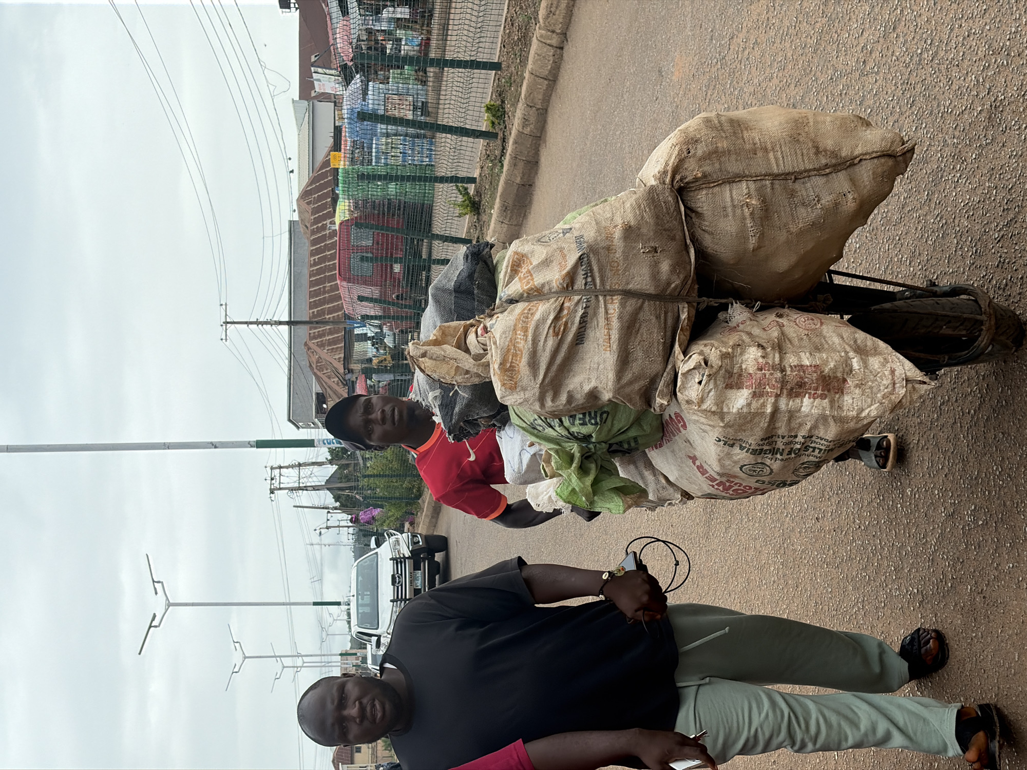 This is a medium shot, outdoor, daytime photograph taken in Bukuru, Nigeria. The scene depicts two men on a dusty road, with market stalls and buildings visible in the background.
In the foreground, a man with dark skin, wearing a black t-shirt and light-colored sweatpants, walks from left to right. He is holding a smartphone and a coiled black cable in his right hand. He is looking forward and to his left. His left hand is by his side. He is wearing black sandals.
To his right, another man with dark skin, wearing a red t-shirt with an orange collar and a black baseball cap, is pushing a cart loaded with large sacks. The sacks appear to be made of burlap and are tied with rope. Some sacks are light brown, and one is dark grey. One of the light brown sacks has text on it that reads "MILLERY PLC" and "NIGERIA". The man is looking forward and to his right. He is wearing dark sandals.
Behind the men, a white Toyota Hilux pickup truck is parked on the road. Further back, there are various market stalls with colorful goods, a red van, and buildings with corrugated iron roofs. Electrical wires are strung across the sky. Streetlights with multiple arms are also visible. The ground is paved with asphalt and gravel. The sky is overcast.