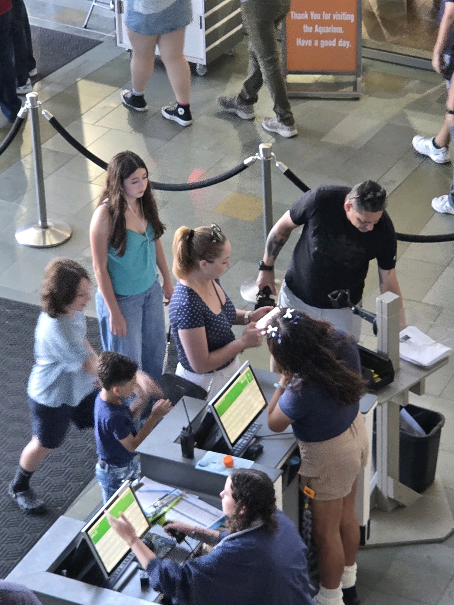 The image depicts an indoor scene, likely within an aquarium or similar public attraction in Long Beach, United States of America, judging by the "Thank You for visiting the Aquarium. Have a good day." sign visible in the upper right corner.
Several individuals are present. In the foreground, a person in a blue shirt and denim shorts is looking at a computer monitor, with their right hand extended towards it. Next to them, a person with long, dark curly hair, wearing a blue top and tan shorts, is facing away from the camera. In the middle ground, a woman with blonde hair in a ponytail, wearing a navy blue polka dot shirt, is looking at a smartphone. To her left, a young woman with long dark hair, wearing a teal tank top and jeans, stands looking down. A young boy in a blue t-shirt and jeans is standing to the left of the polka dot shirt woman, looking at a computer monitor. Further back, a man in a black t-shirt and grey shorts is looking down at something in his hands, with tattoos visible on his left arm. Another person, partially visible, is walking in the background.
The environment includes a grey counter with multiple computer monitors, a scanner, and a black trash can. Stanchions with black ropes are also visible, indicating a queuing area or pathway. The floor is tiled. The lighting appears to be artificial, casting some shadows.