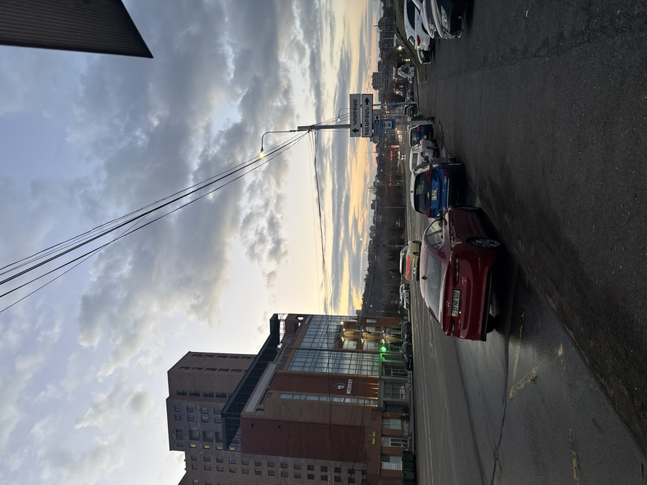 The image depicts an urban street scene at dusk on M. Baghramyan Avenue 21, Yerevan, Armenia, near the Medesy Medical Center. The foreground features a wet asphalt road with yellow markings on the left, and a paved sidewalk on the right. A red sedan (Armenian license plate 37 NR 037) is parked curbside on the left, partially on the road. Behind it, on the sidewalk, a blue Chevrolet Bolt EV and several other vehicles are parked in a line.
In the midground, a modern multi-story building with a red-brick and glass facade, displaying the "MEDESY" logo and "21 BAGRAMYAN" signage, dominates the left side. A taller, older-style brown residential building stands behind it. On the right, a utility pole with an illuminated streetlamp supports multiple overhead power lines and street signs in Armenian script, including "ԿՈՄԻՏԱՍ" (Komitas) and "ՀԱՆՐԱՊԵՏՈՒԹՅԱՆ" (Hanrapetutyan), indicating directions. Moving vehicular traffic, including a white sedan with illuminated taillights, is visible on the main road. The background reveals a distant hilly landscape with buildings and sparse vegetation under a cloudy sky, transitioning to an orange and yellow sunset on the horizon. No individuals are clearly visible in the frame.