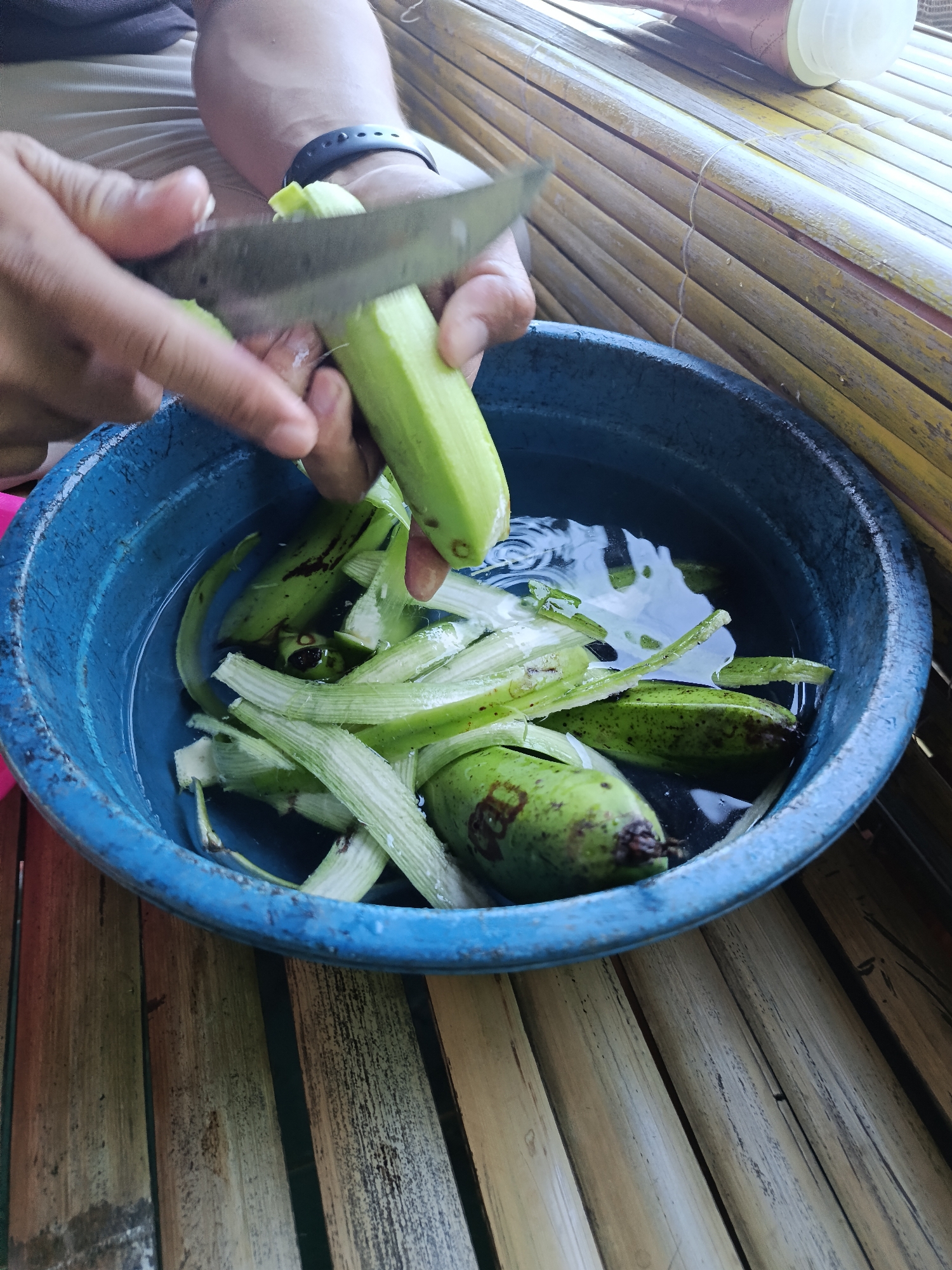 In Mansalean, Indonesia, a person is preparing green bananas in a blue plastic basin filled with water. The person's hands, one holding a knife, are actively peeling the bananas. Several peeled banana skins, long and fibrous, are submerged in the water along with the unpeeled and partially peeled bananas. The basin rests on a surface of bamboo. The surrounding environment appears to be outdoors or in an open-air setting, suggested by the bamboo structure.