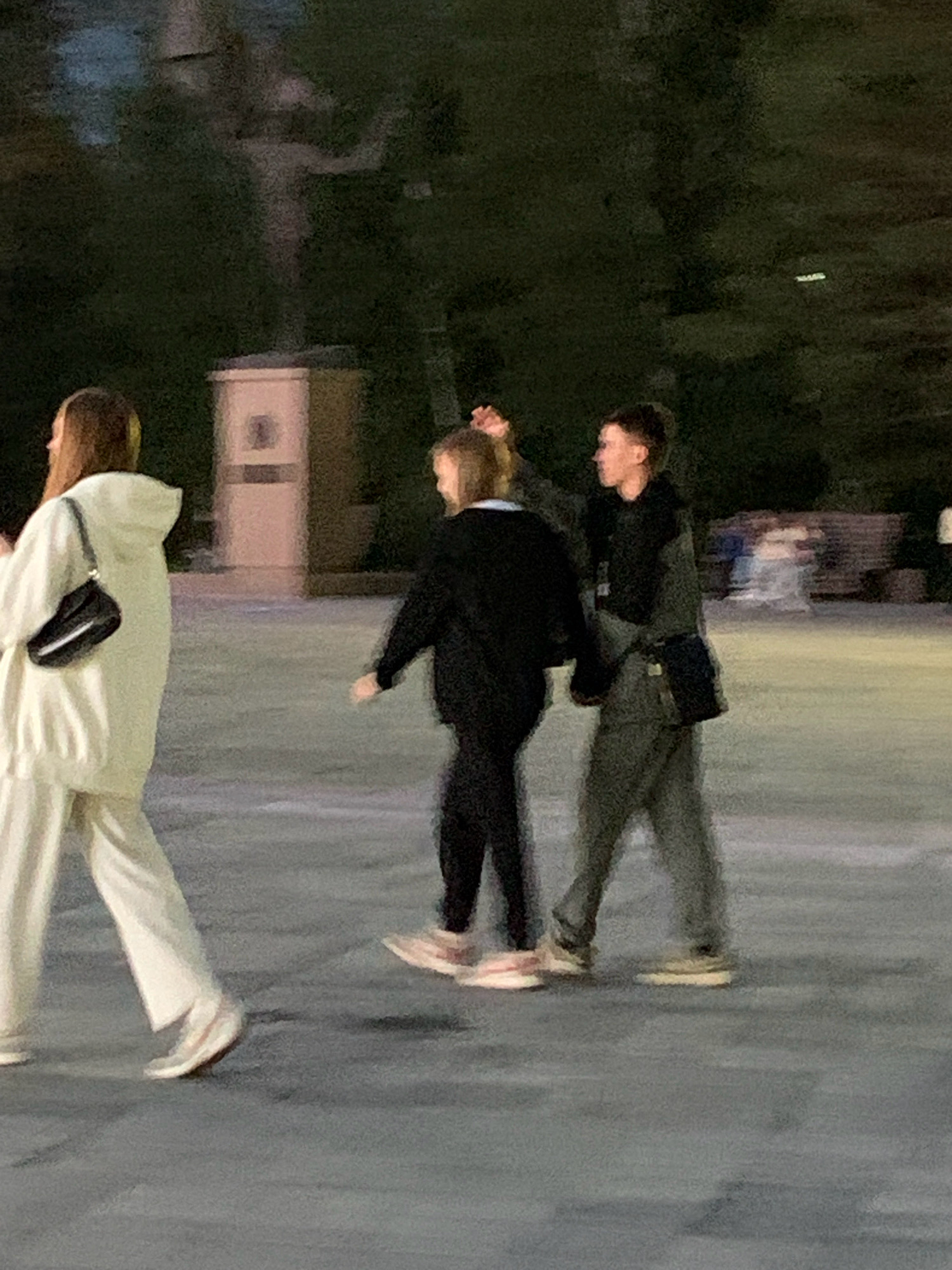 An outdoor, nighttime, slightly blurry image shows three people walking on a paved surface. On the far left, a person in a light-colored hooded tracksuit and white sneakers walks away from the camera. A black purse is slung across their chest. In the center, a person in a black hoodie and black pants, with pink and white sneakers, walks towards the right, looking back over their shoulder. A person in a gray tracksuit and light-colored sneakers walks to the right of the center person, with a black bag slung across their chest, and their right hand is raised, touching the back of the center person's head. In the background, a tall, light-colored, square structure with a circular emblem is visible, as well as dark trees and blurred lights. The scene appears to be an urban setting at night, possibly a park or public square, in Prokopyevsk, Russia.