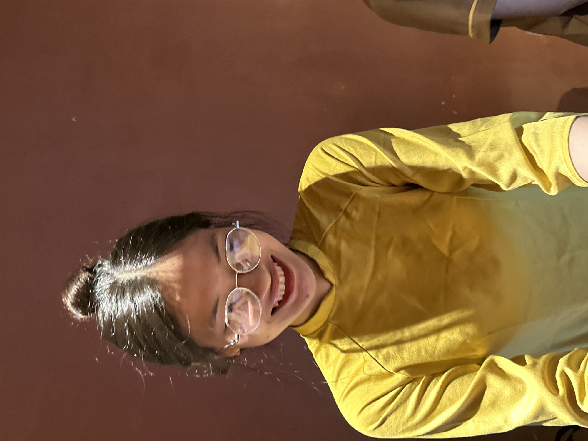 A young woman with dark hair pulled into a top knot and wearing round, silver-rimmed glasses smiles broadly at the camera. She is dressed in a long-sleeved yellow shirt with a subtle gradient, lighter at the bottom. The background is a plain, warm brown wall. The image appears to have been taken indoors, possibly in a cafe or restaurant, in Đà Nẵng, Vietnam. There is a faint reflection visible in her glasses.