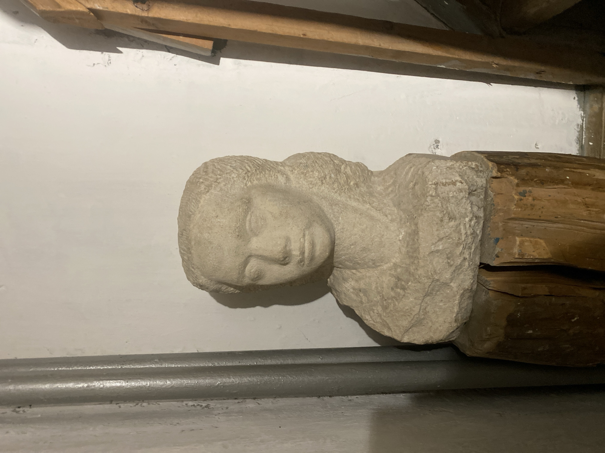 The image shows a stone bust of a woman with braided hair, resting on a rough-hewn wooden pedestal. The bust is made of a light-colored stone and depicts the woman from the shoulders up, with her head slightly tilted. Her facial features are detailed but appear weathered, and her eyes are closed. The wooden pedestal is made of several pieces of wood, showing signs of age and wear.
The bust is positioned indoors, against a white wall. To the left of the bust is a vertical, ribbed metallic element, possibly a pipe or part of a column. To the right of the bust, a wooden structure is partially visible, appearing to be a vertical beam or support. The lighting in the scene is artificial, casting shadows that highlight the textures of the stone and wood.
The image does not provide any information about the location within the city of Moscow, Russia.