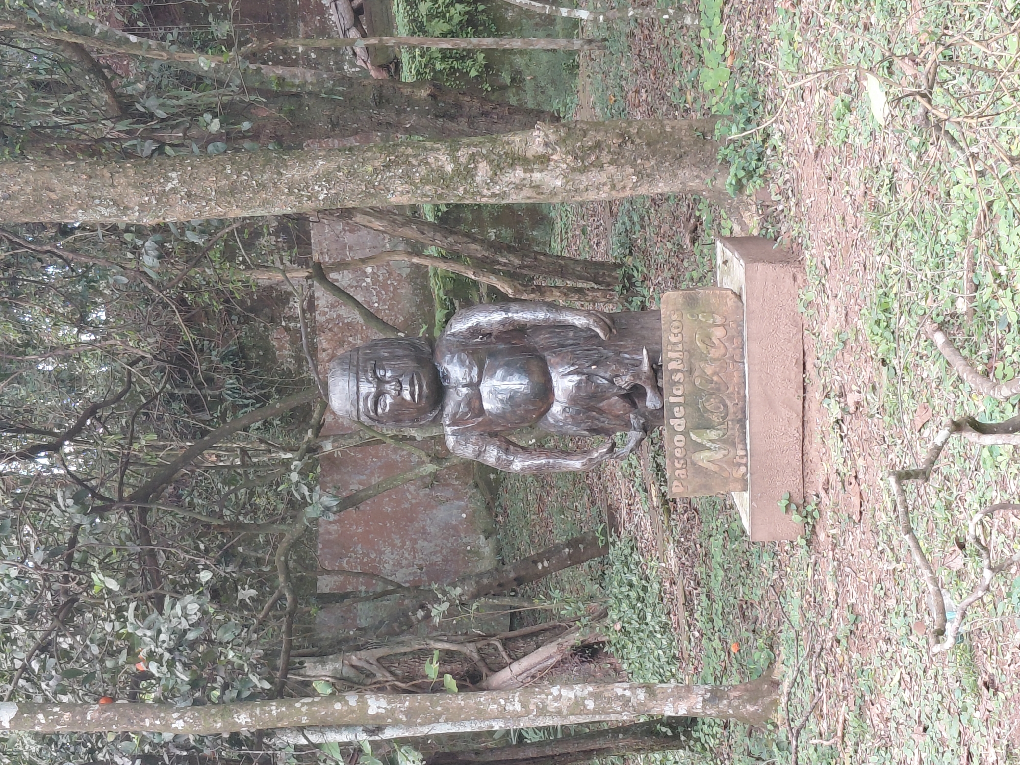 The image shows a metallic statue of a person in a seated position, with a stern expression and muscular physique, positioned on a stone pedestal in a wooded outdoor setting. The pedestal has text carved into it, reading "Paseo de los Mitos" and "Morroi". The statue is dark in color and appears to be made of weathered metal. The surrounding environment consists of trees with visible bark and foliage, and the ground is covered with fallen leaves and green vegetation. The background shows a rustic wall or structure. The scene is likely in a park or historical site, given the nature of the statue and its inscription. The city of Trinidad, Paraguay, is known for its Jesuit missions and associated historical sites, making it a plausible location for such a monument.