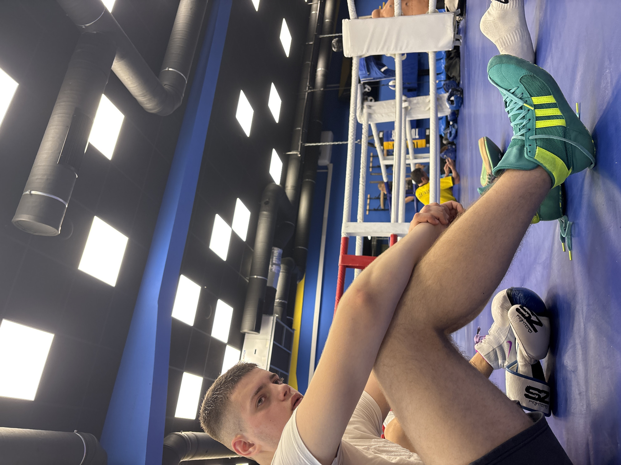 This image captures an indoor scene within a boxing or martial arts gym, potentially located in Sofia, Bulgaria. The main subject is a young male, seen from a low angle, resting on a blue mat surface. He has short, light-colored hair and is wearing a white t-shirt and dark shorts, with bare legs. His right leg is bent with his foot on the mat, while his left leg is extended upwards, bent at the knee, with his hands clasped around it. He gazes upwards and slightly to his right, with a calm, contemplative expression.
On his left foot, he wears striking teal-green Adidas boxing boots featuring bright yellow three-stripe branding and matching yellow accents. White socks are visible above the boots. Beside his right leg, a pair of white and blue boxing gloves rests on the mat, clearly displaying the brand "SZ FIGHTERS" on their cuffs.
The background reveals a white-roped boxing ring with blue corner posts and some red rope trim, indicating a professional training setup. Other individuals are present: one person is seen sitting on the edge of the boxing ring, and further back, another person wearing a yellow top is visible inside or near the ring. The gym features blue walls and a dark grey/black ceiling. The ceiling is equipped with numerous bright, square and rectangular recessed lights that illuminate the space, and large black ventilation ducts run across the ceiling. The setting conveys an active training environment, well-lit by artificial lighting.