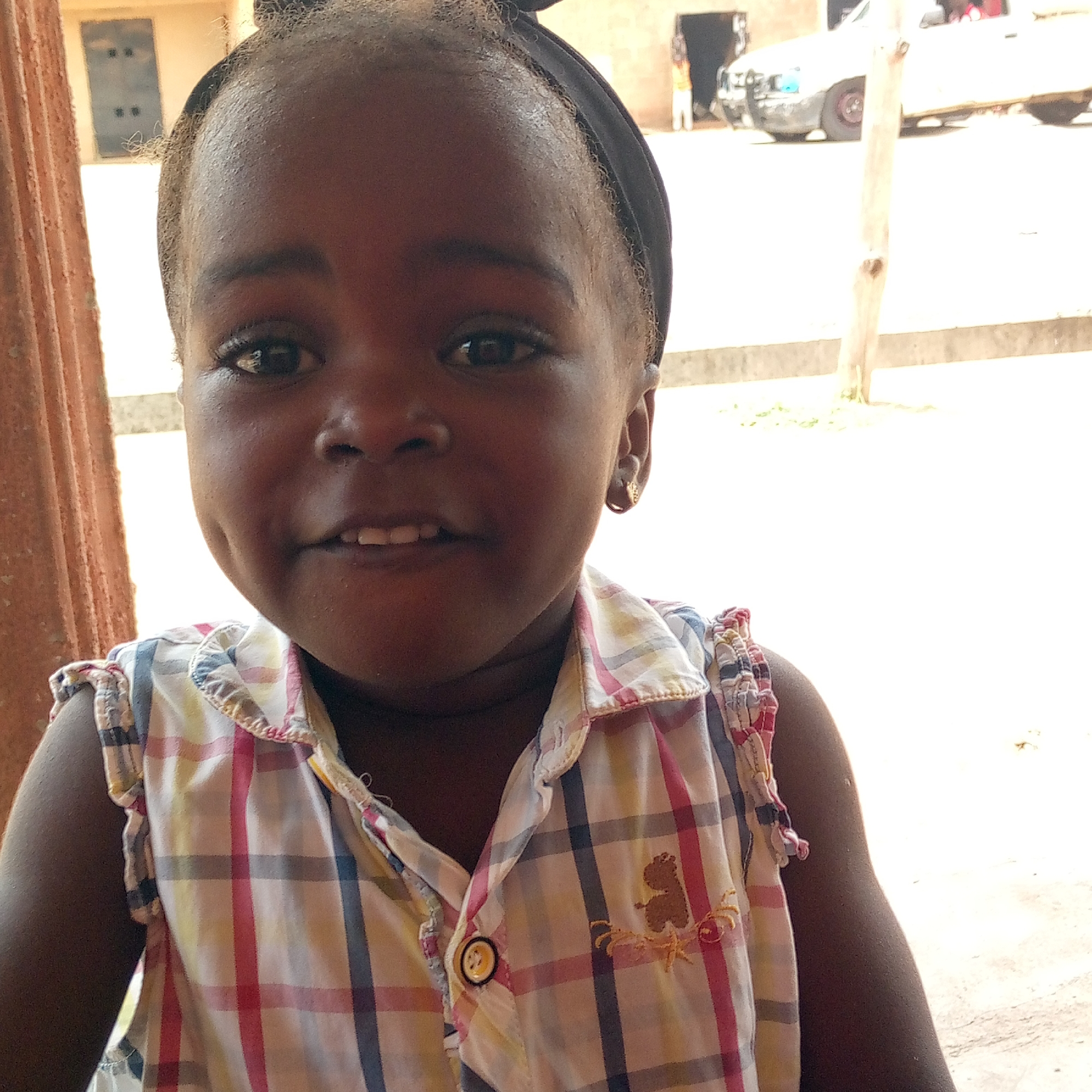 A young African girl with a black headband smiles at the camera. She wears a plaid shirt with a bear design and a button. The background includes a concrete pillar to the left, a building with a window, and a white pickup truck on a street. The location appears to be outdoors in daylight. The environment is potentially a street-side area in Mararaba, Nigeria, based on the prompt's information.