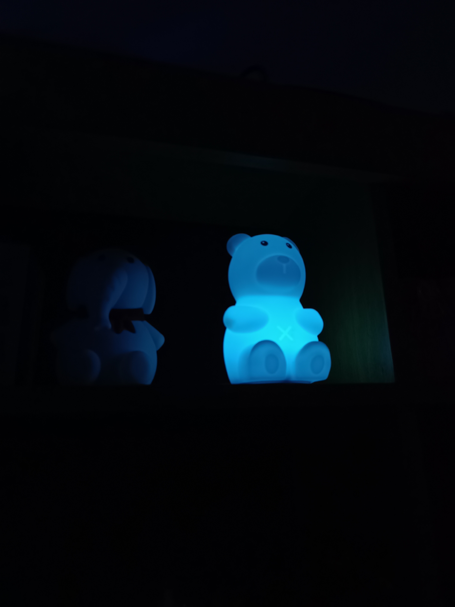 The image shows two illuminated, cartoon-like toy figures positioned on a dark shelf. The figure on the left appears to be an elephant-like shape with a small bow around its neck. The figure on the right is a teddy bear shape with an "X" on its chest. Both figures emit a blue-toned light, which is the primary source of illumination in the scene. The background is dark, revealing only the shelf's surface on which the figures stand. Location is not evident, Cherkovitsa, Bulgaria cannot be confirmed or denied as the image offers no context.