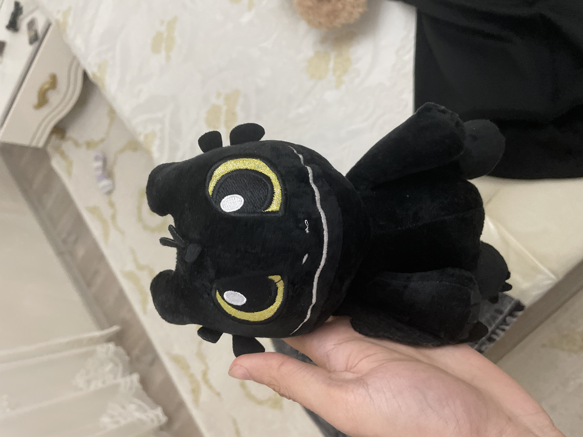 A close-up image, slightly rotated, features a human hand holding a black plush toy against an indoor background. The hand, with light skin, gently supports the toy in the lower half of the frame.
The plush toy is distinctly black, made from a soft, presumably velour-like material. It has prominent, large, and expressive embroidered eyes that are bright yellow with a glittery finish, dark pupils, and small white highlights. Small, rounded ear-like protrusions extend from the top of its head, and delicate white stitching lines adorn its face and body, adding subtle detail. The toy appears to be a stylized creature, possibly a dragon or a similar mythical animal.
The background shows a light-colored surface, likely a bedspread or sofa cover, with a subtle pattern of pale yellow or beige leaf-like motifs on a white or off-white fabric. To the left, a portion of light-colored furniture, possibly a bedside table or dresser, is visible, featuring elegant curves and a small, gold-colored decorative accent. In the upper right background, a dark, indistinct object, potentially another piece of fabric or a larger plush item, adds to the domestic setting.
The lighting in the image is soft and even, suggesting an indoor environment, possibly illuminated by indirect natural light or artificial lighting. No text is visible on the toy or in the background. The overall scene conveys a sense of comfort and a personal space, likely within a home in Ihtiman, Bulgaria.