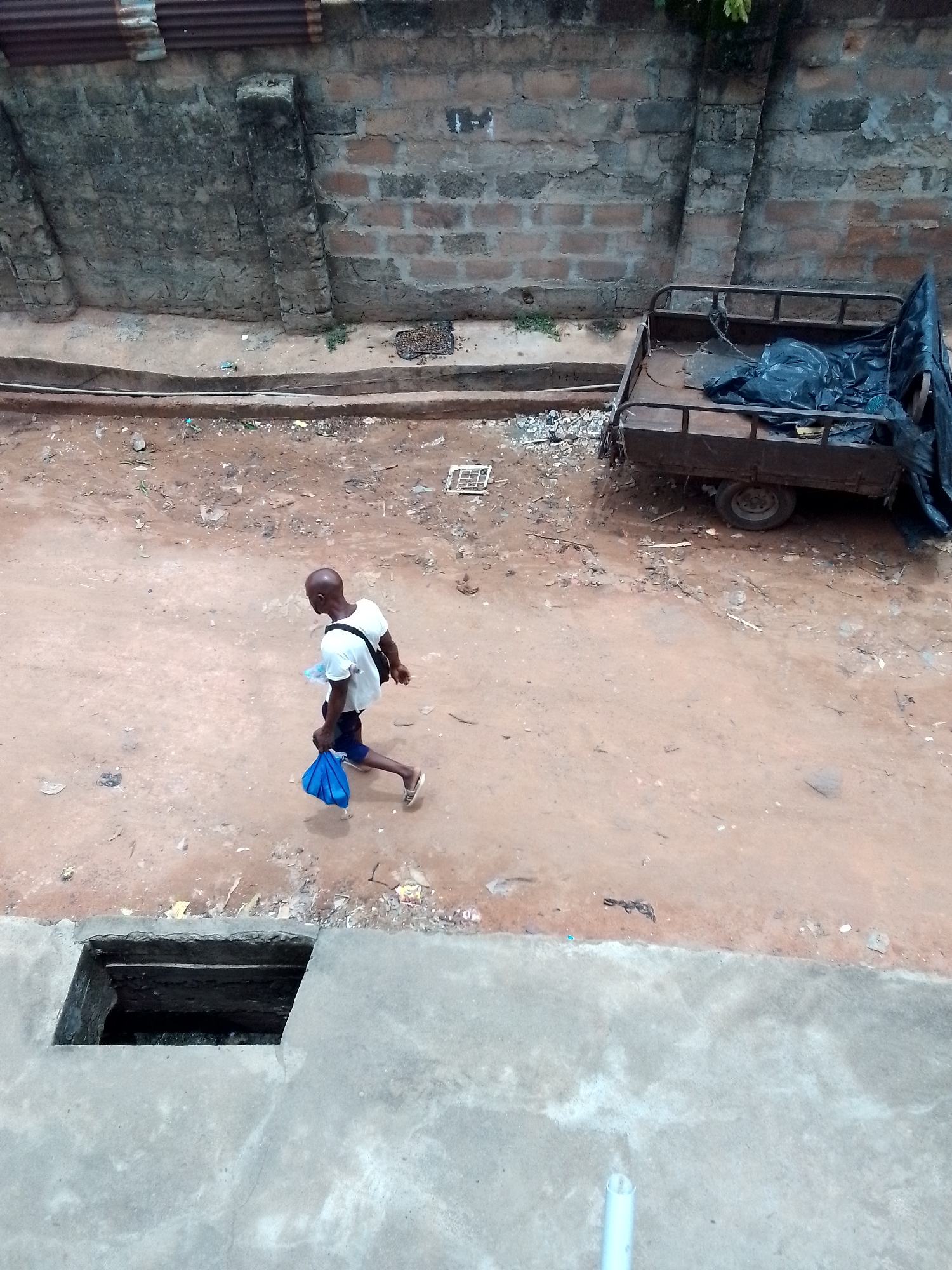 A high-angle shot reveals a street scene in Unknown, Unknown. A bald man, wearing a white t-shirt and dark shorts, walks across a dirt road, carrying a blue bag. He has a dark shoulder bag. To his right is a trailer, covered with a black tarp. In the lower left, a square hole is present in the concrete ground. A brick wall stands behind the road. The ground is dirt with scattered debris.