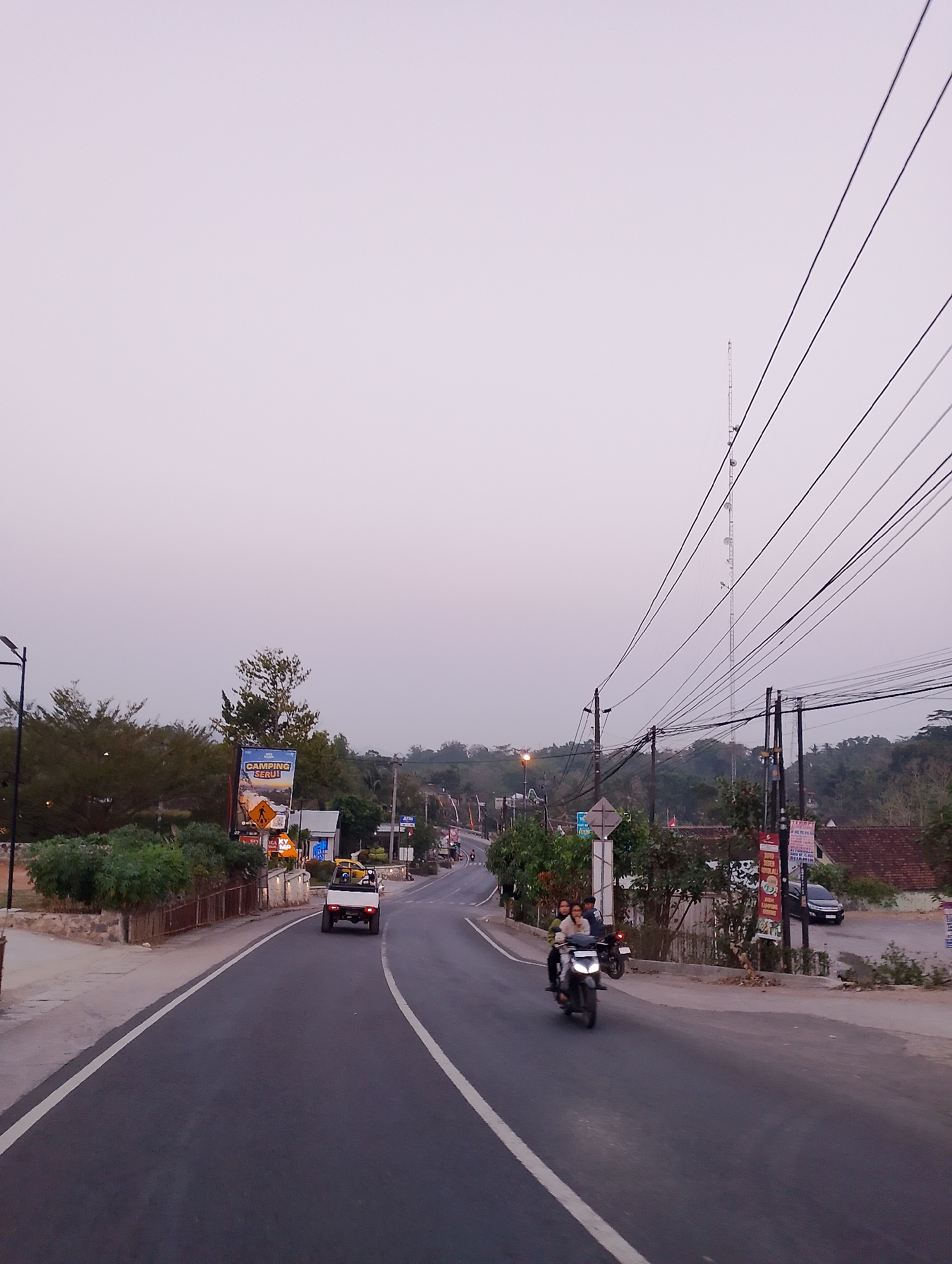 The image depicts a road in Patuk, Indonesia, during twilight. A paved road curves through a rural landscape with trees and rolling hills in the background. Several utility poles with numerous wires are visible along the roadside, along with a tall communication tower in the distance. On the road, a white open-back vehicle is driving away from the viewer, and a motorcycle carrying two passengers is approaching. Various signs, including advertisements and traffic signs, are present. Buildings, some with red-tiled roofs, are interspersed with the vegetation. The sky is overcast and light purple, indicating the end of daylight.