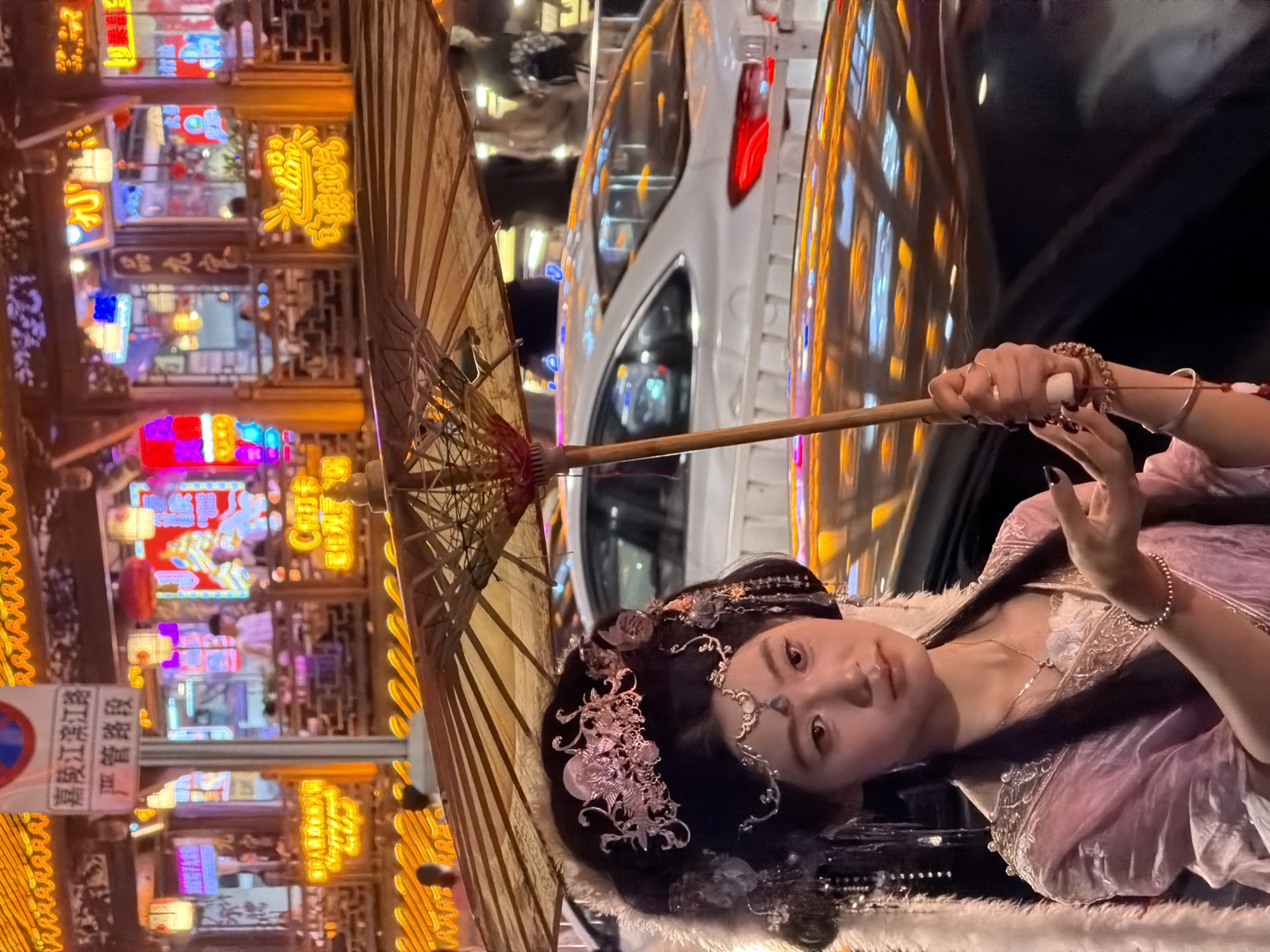A young East Asian woman wearing a pink, ornate traditional dress and an elaborate hair ornament stands holding a traditional umbrella. The umbrella is open and angled towards the viewer, obscuring the upper part of the woman's face and the background. She has long, dark hair, with strands falling around her shoulders. Her makeup is subtle, with emphasis on her lips. She is wearing a delicate necklace and bracelets on her wrists. The background is a bustling street scene at night, illuminated by neon lights and glowing signs. Buildings with traditional architectural elements are visible, adorned with vibrant, colorful lights. The reflection of these lights can be seen on the surface of a car parked behind the woman and on the umbrella itself. A white car is parked behind her, and its tail light is illuminated. The overall scene depicts a lively urban environment at night, possibly in the Yuzhong District of China, characterized by bright lights and traditional aesthetics.