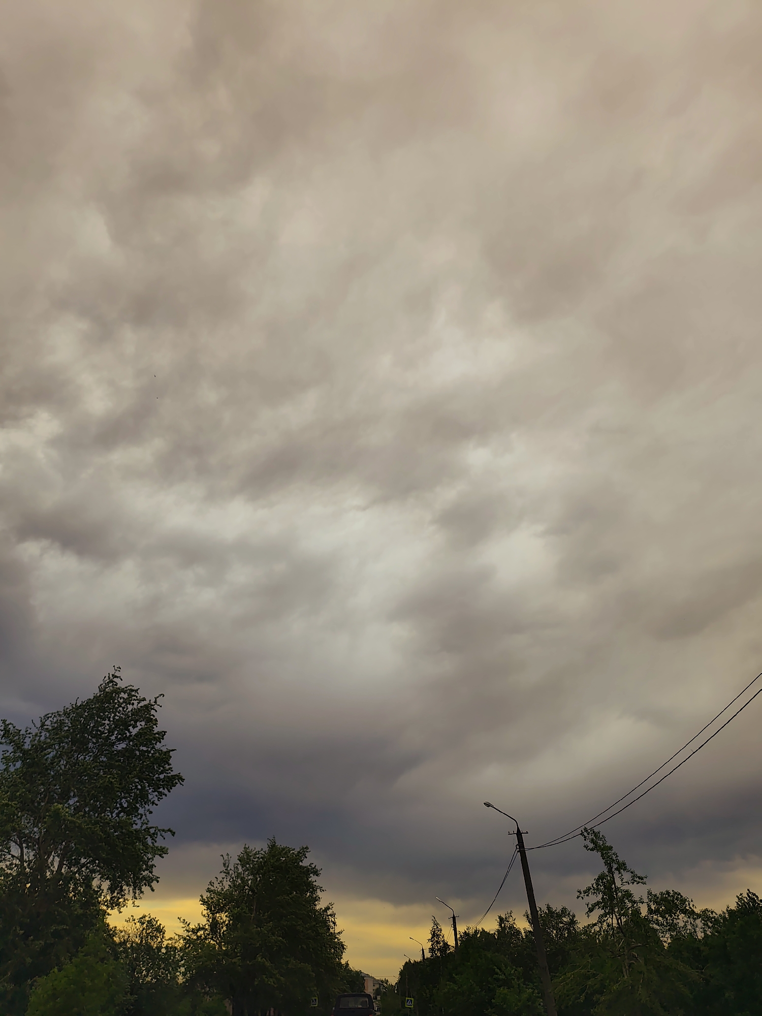 The image depicts an overcast sky above a street in Gremyachinsk, Russia. The sky is filled with thick, gray clouds, suggesting an impending storm. Below, trees and foliage line the street, partially obscuring the view of the road. Utility poles and power lines are visible against the cloudy sky. A warm, golden light is visible below the clouds on the horizon.