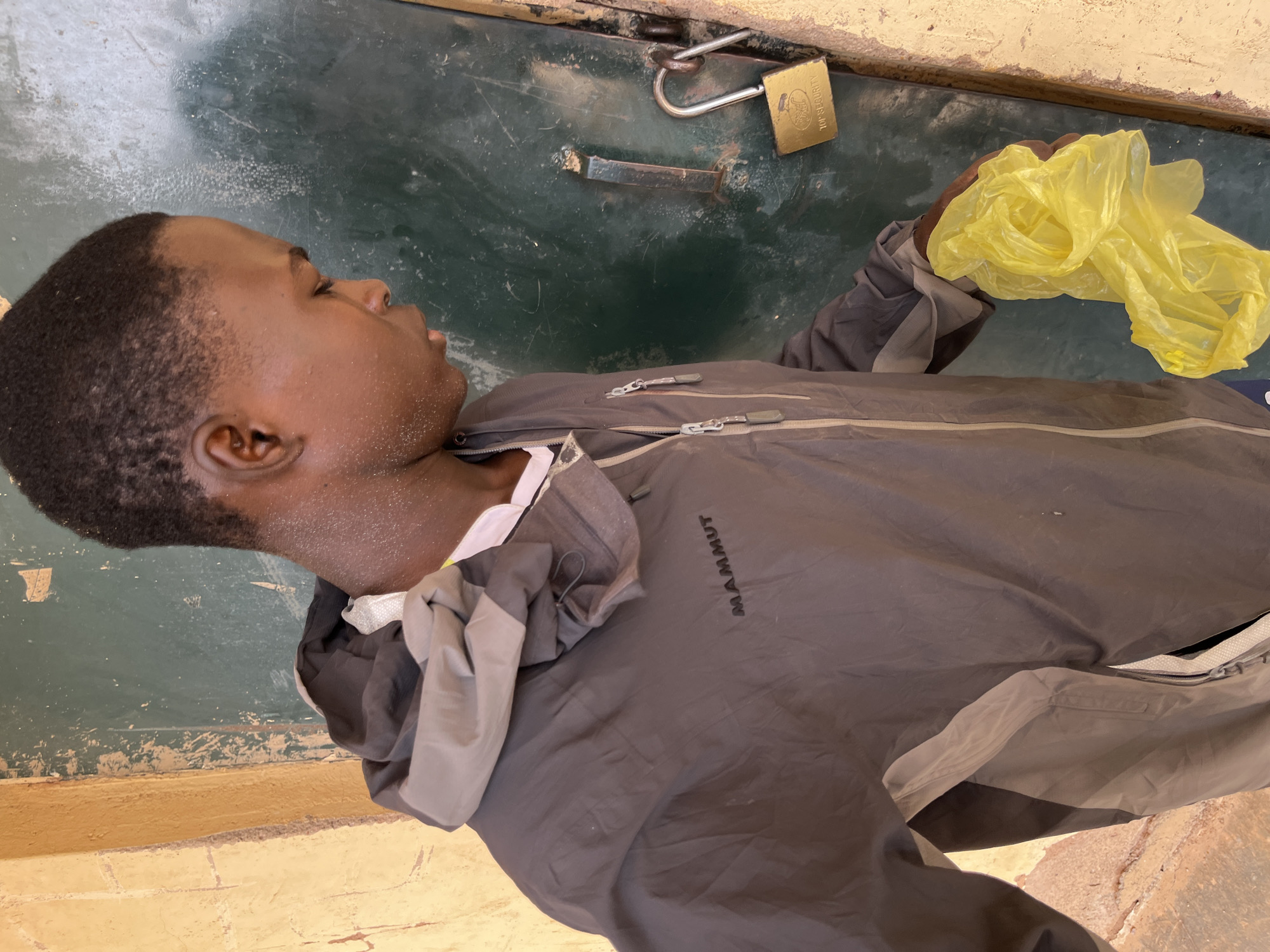 In Bukuru, Nigeria, a young boy in a grey and black hooded jacket is standing in front of a green metal door. The door is secured with a brass padlock that reads "TOP SECURITY". The boy is holding a crumpled yellow plastic bag in his right hand. The wall to the left of the door is a rough, sandy texture, and the door frame appears to be made of wood. The scene suggests a mundane moment, possibly of the boy arriving at or departing from a locked premises.