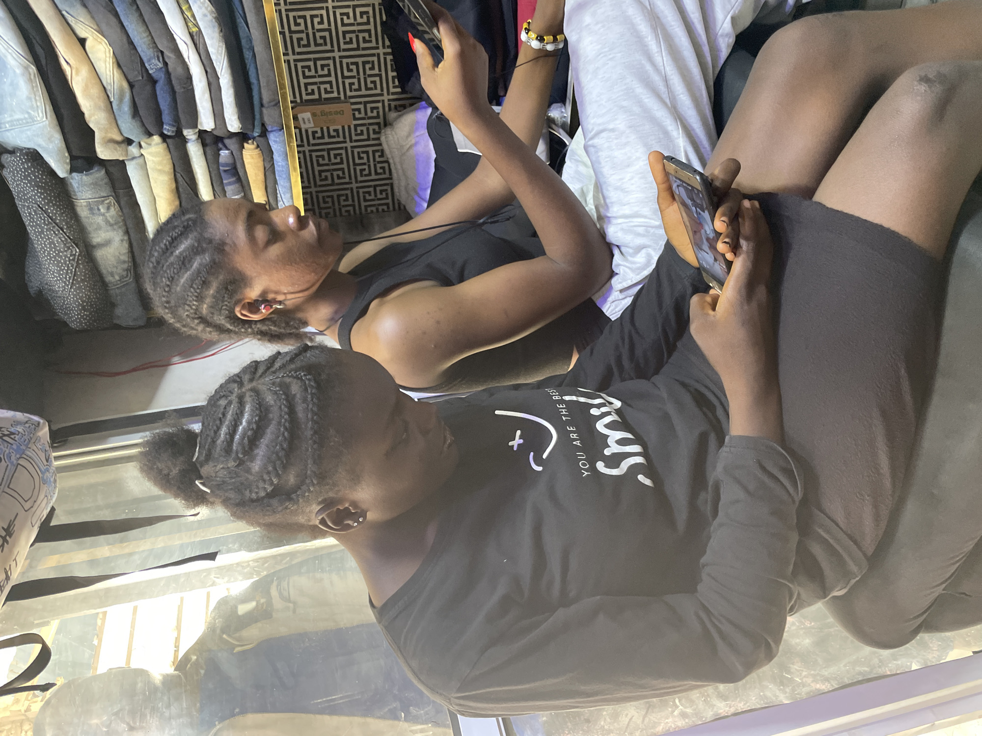 Two young women with braided hair are seated side-by-side, each focused on their mobile phones. One is wearing a black tank top, while the other wears a long-sleeved black shirt with white text that says "smile". Clothes on hangers can be seen behind one of the women, suggesting they may be inside a store or market. The photo was taken in Bukuru, Nigeria.