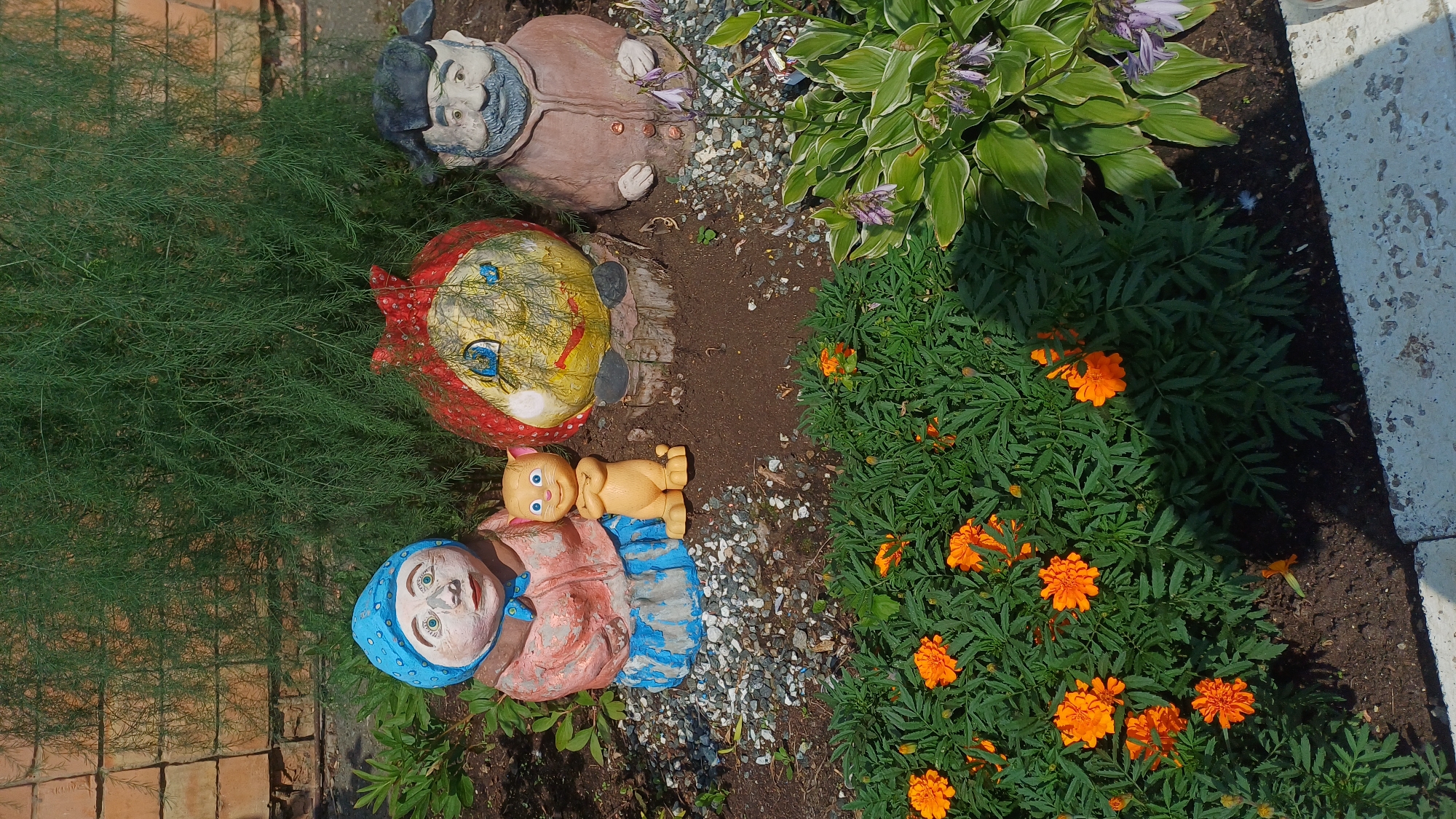 A vertical, medium shot captures a garden scene with three decorative figures and blooming flowers. The figures, positioned in the upper center, are a woman in a blue headscarf and pink coat, a yellow cat figurine, a round, yellow character with a red headscarf, and a man with a black hat and brown coat. The figures are placed on dark soil and gravel. In the foreground, to the left, are bright orange marigolds. To the right, there are hosta plants with pale purple flowers. Behind the figures, a tall, feathery green plant stands against a brick wall. The scene is outdoors on a sunny day. This image was taken in Izhevsk, Russia.
