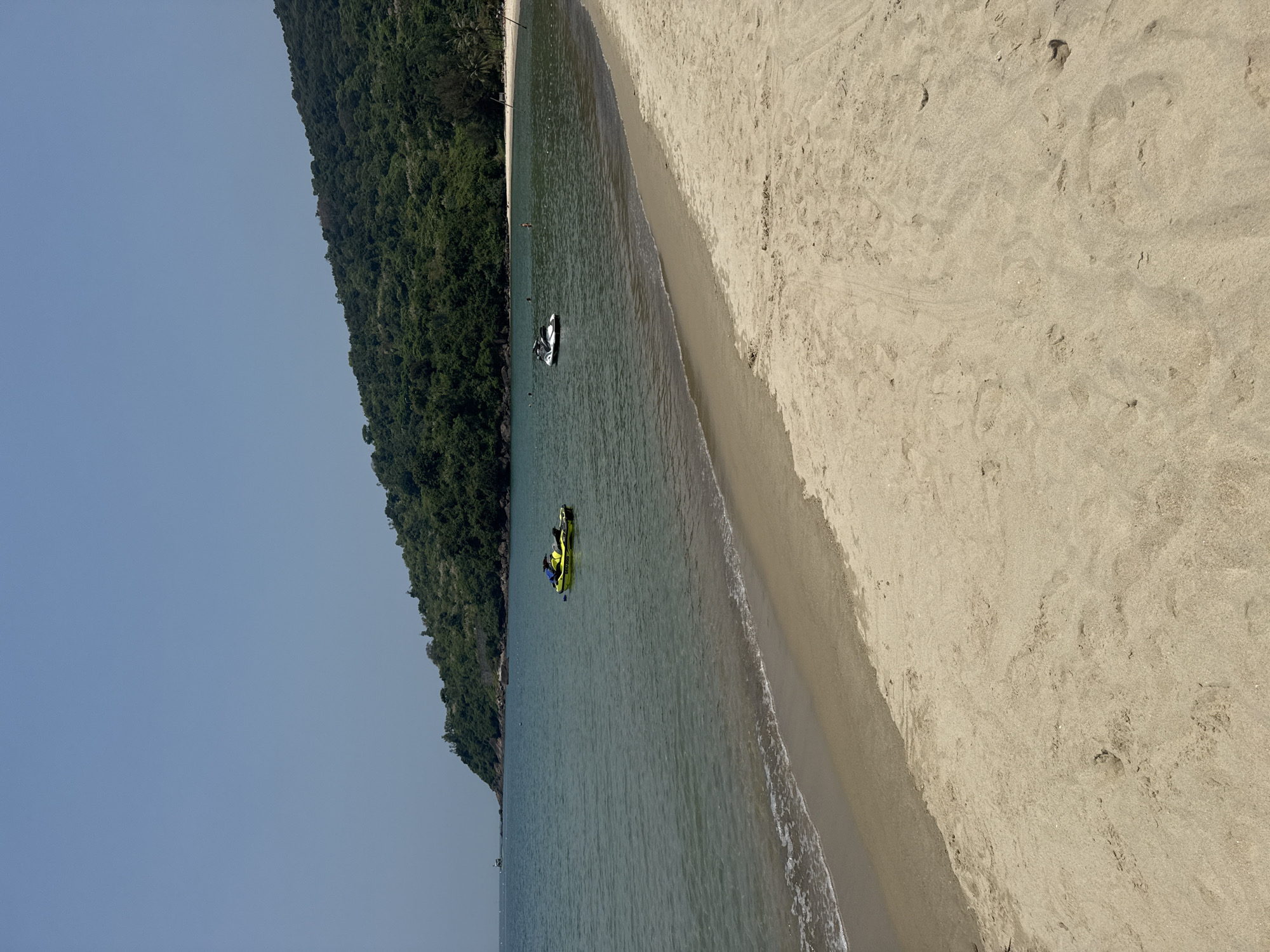 The image captures a daytime coastal scene on the Sơn Trà Peninsula, Đà Nẵng, Vietnam. The foreground features a light brown sandy beach exhibiting various foot impressions, transitioning to wet sand at the waterline where small waves recede. The midground shows a calm bay with clear, greenish-blue water. Two personal watercraft are visible, a yellow-green model positioned on the left side of the bay and a white-black model further to the right. Several indistinct individuals are observed in the water, appearing to be swimming or wading. A line of white buoys stretches across the bay, delineating a section of the water. A distant white vessel is discernible on the far left horizon. The background is dominated by a steep, densely forested mountain covered in dark green foliage, rising directly from the water's edge and extending across the right half of the frame. The sky above is clear and light blue.