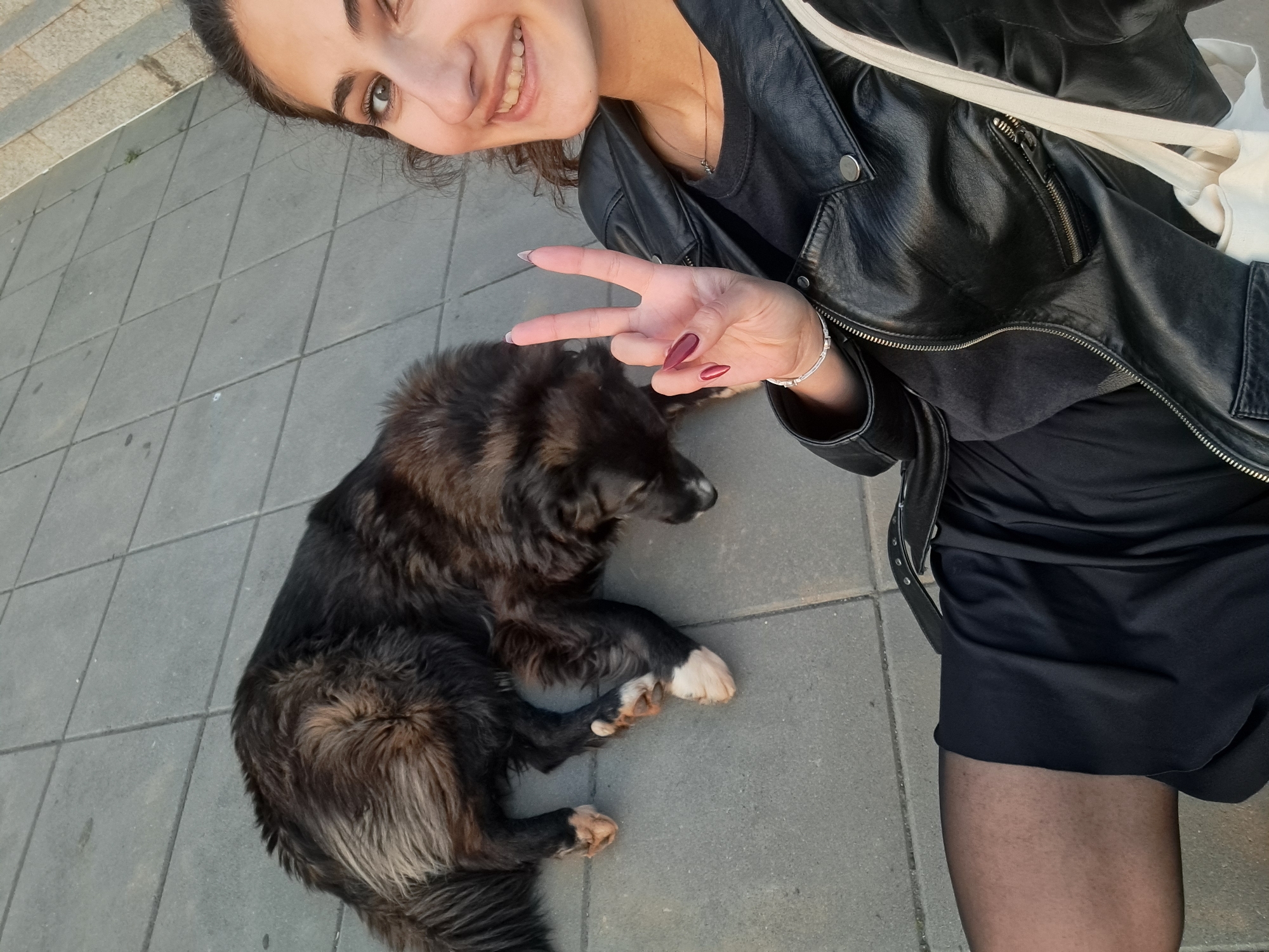 A woman in a black leather jacket and black dress kneels on a gray tiled surface next to a medium-sized, dark brown dog with white paws. The woman is smiling and making a peace sign with her right hand. A white tote bag is draped over her shoulder. The scene is set outdoors, possibly a sidewalk or plaza. The exact location in Negovan, Bulgaria is not discernible from the image.
