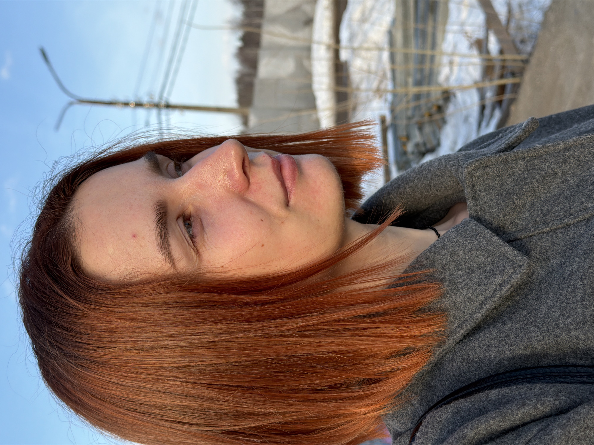 A sideways outdoor shot depicts a fair-skinned woman with reddish-brown hair wearing a gray wool coat. She is looking upwards and slightly to the right. The background shows a blurry cityscape with a street lamp and indistinct buildings. The location is Chaikovsky, Russia, suggested by possible background objects.