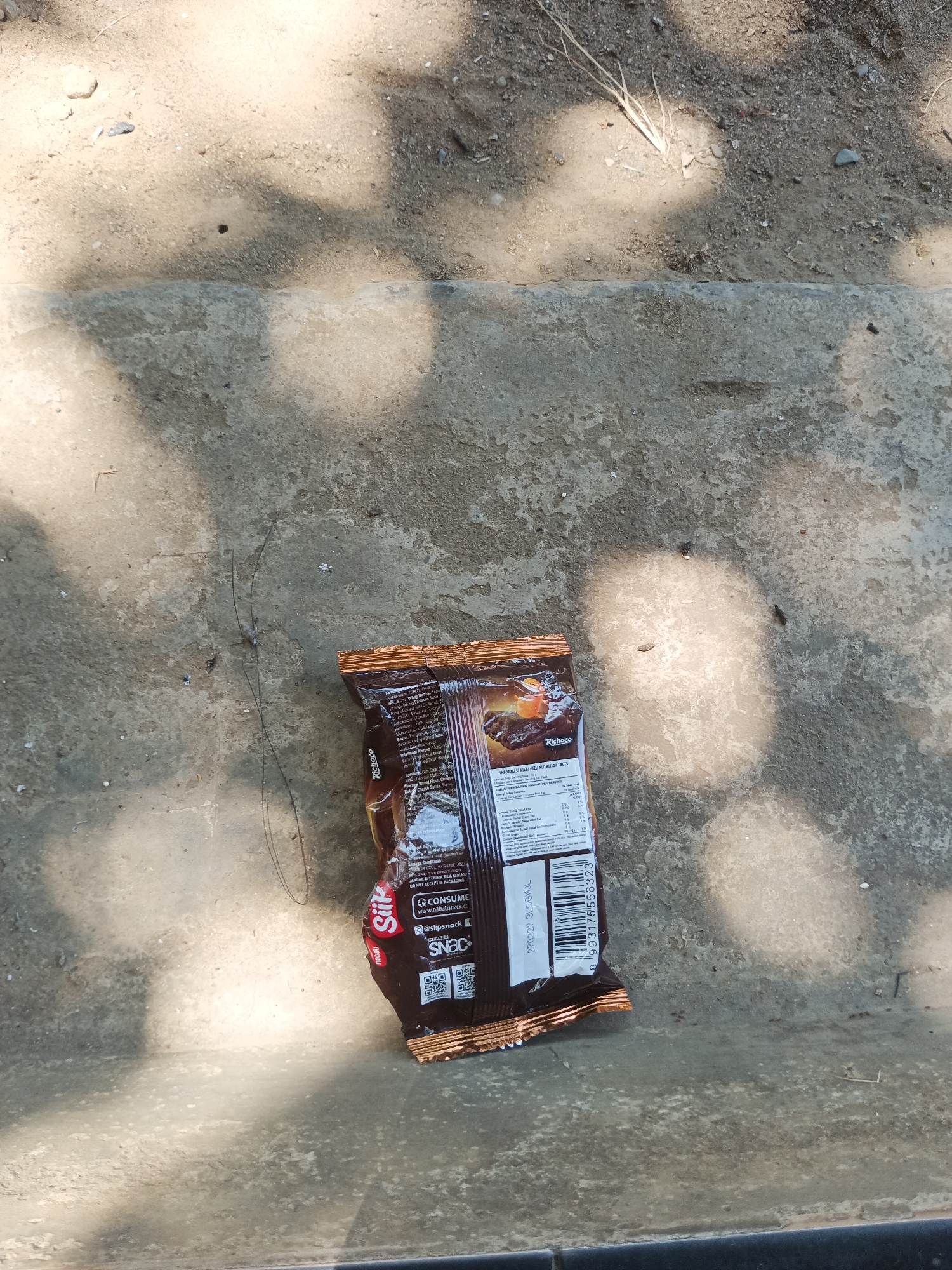 A single, opened packet of Richoco Siip wafer is lying on a concrete surface outdoors, possibly a set of steps or a walkway. The packet is dark brown with orange and brown text and images on it. The text on the packet is mostly illegible due to the image resolution and the condition of the packet, but some readable elements include "Richoco," "Siip," "INFORMASI NILAI GIZI NUTRITION FACTS," and a barcode. There are shadows from overhead foliage cast on the concrete, suggesting it is daytime. The surrounding environment appears to be sandy and dusty, with some small pebbles and debris. There are no people visible in the image. The scene is consistent with a street or public area in Pitue, Indonesia.