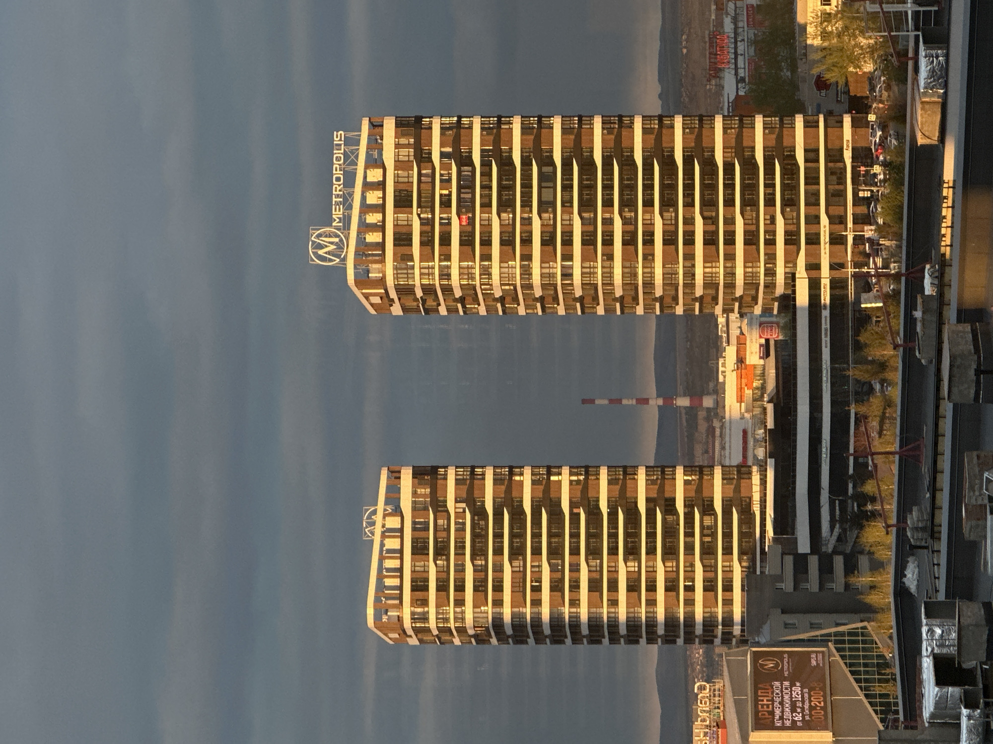 The image shows two high-rise buildings in Krasnoyarsk, Russia, labeled "Metropolis" on top. They are situated in an urban environment with other buildings, construction materials, and a billboard visible. The background includes what appears to be a hazy or overcast sky. The billboard advertises commercial real estate for rent in the Metropolis complex at 16 Oktyabrskaya Street. A red-and-white striped industrial chimney is visible in the distance between the two buildings.
