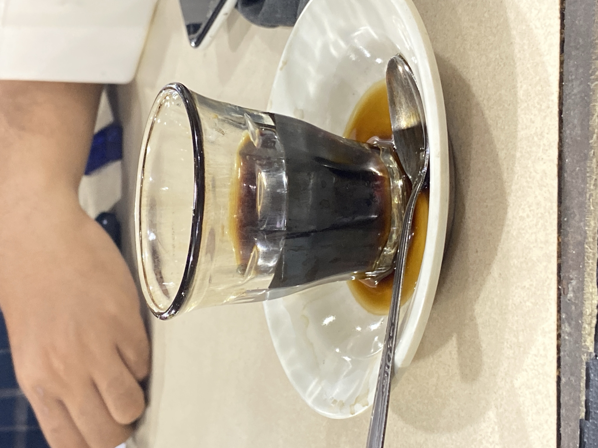The image shows a close-up, top-down view of a small glass of dark liquid, likely coffee or tea, sitting on a saucer. A spoon rests on the saucer, partially submerged in some of the spilled liquid. The glass has a slightly flared rim and is made of clear glass, allowing the dark beverage to be seen. There are a few drops of the liquid on the white saucer, indicating it may have been recently moved or spilled.
To the left of the glass and saucer, a person's hand and arm are visible. The person is wearing a blue and white striped garment. The background is a light-colored, textured surface, possibly a table, with a dark vertical line on the right edge suggesting a separation or edge of the table.
The setting appears to be indoors, possibly a cafe or restaurant, given the presence of a beverage served in a glass and saucer. The lighting suggests it is daytime. No text is visible in the image. The overall impression is a casual moment captured while someone is enjoying a drink.