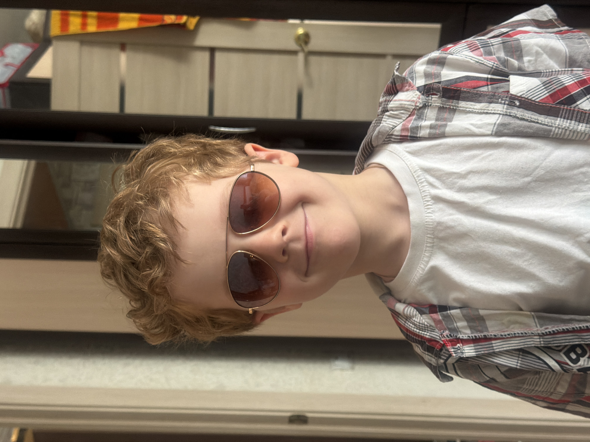 A young, fair-skinned child with curly blonde hair wears rose-tinted aviator sunglasses. The child is wearing a white t-shirt covered by a gray, white, and red plaid flannel shirt. The background includes a dresser with drawers and a brass knob on one of the drawers. The photo was taken indoors. I cannot confirm the location in Chaikovsky, Russia.