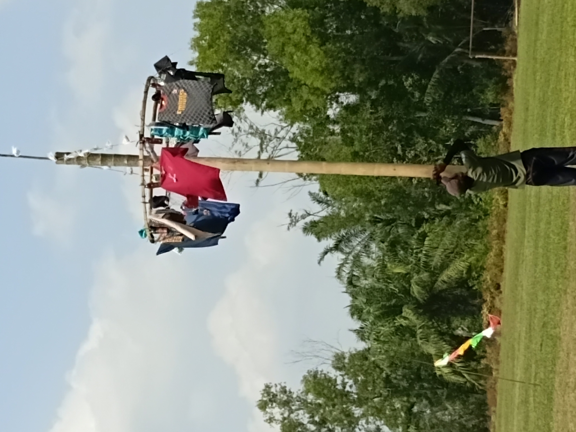 The image depicts a tall, wooden pole structure adorned with various articles of clothing, resembling a makeshift clothesline or decoration. The pole is situated in an open, grassy field, with a line of trees forming a backdrop. In the foreground, a person dressed in a green shirt and dark pants is seen looking up at the pole, their arms raised as if interacting with it. A colorful kite is visible in the middle ground to the left of the pole, tethered to a thin stick. The sky above is partly cloudy with a light blue hue. The overall scene suggests an outdoor event or activity taking place in a rural or semi-rural environment, potentially in Margo Rukun, Indonesia, given the presence of tropical vegetation and the general appearance of the landscape.