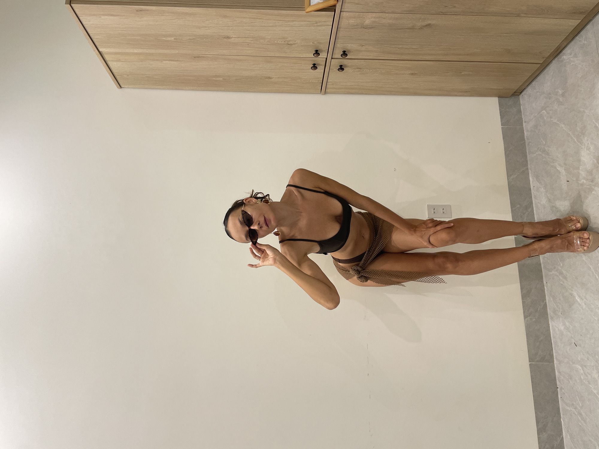 A woman poses indoors, depicted in a full-body shot. She stands angled towards the camera, with her body turned slightly to her right. Her right leg is extended, while her left knee is bent, and her left hand rests on her thigh. She wears dark sunglasses and holds the frame with her right hand near her face. Her dark hair is pulled back. She is dressed in a black bikini top paired with a sheer, light brown or gold mesh sarong tied around her waist. She completes her outfit with light-colored open-toed heels featuring clear straps.
The background consists of a plain, light off-white wall behind and to her left. To her right, a grey tiled floor with a subtle marble-like pattern is visible. In the upper part of the image, light wooden cabinets or wardrobes with dark circular handles span the width, suggesting built-in furniture. A white electrical outlet with two sockets is noticeable on the wall behind her left leg. The scene is brightly and evenly lit, indicative of an indoor setting during daytime. No text is visible in the image. The overall impression is one of confidence and style within a modern interior, located in Mũi Né, Vietnam.