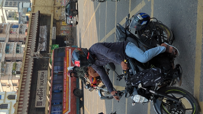 A motorcyclist wearing a helmet and casual clothing is seated on a black motorcycle. Another motorcycle with a rider is visible behind him. The scene takes place in an urban area near buildings and a bus. A second helmet rests on the motorcycle behind the seated rider. Yellow parking stripes mark the road.