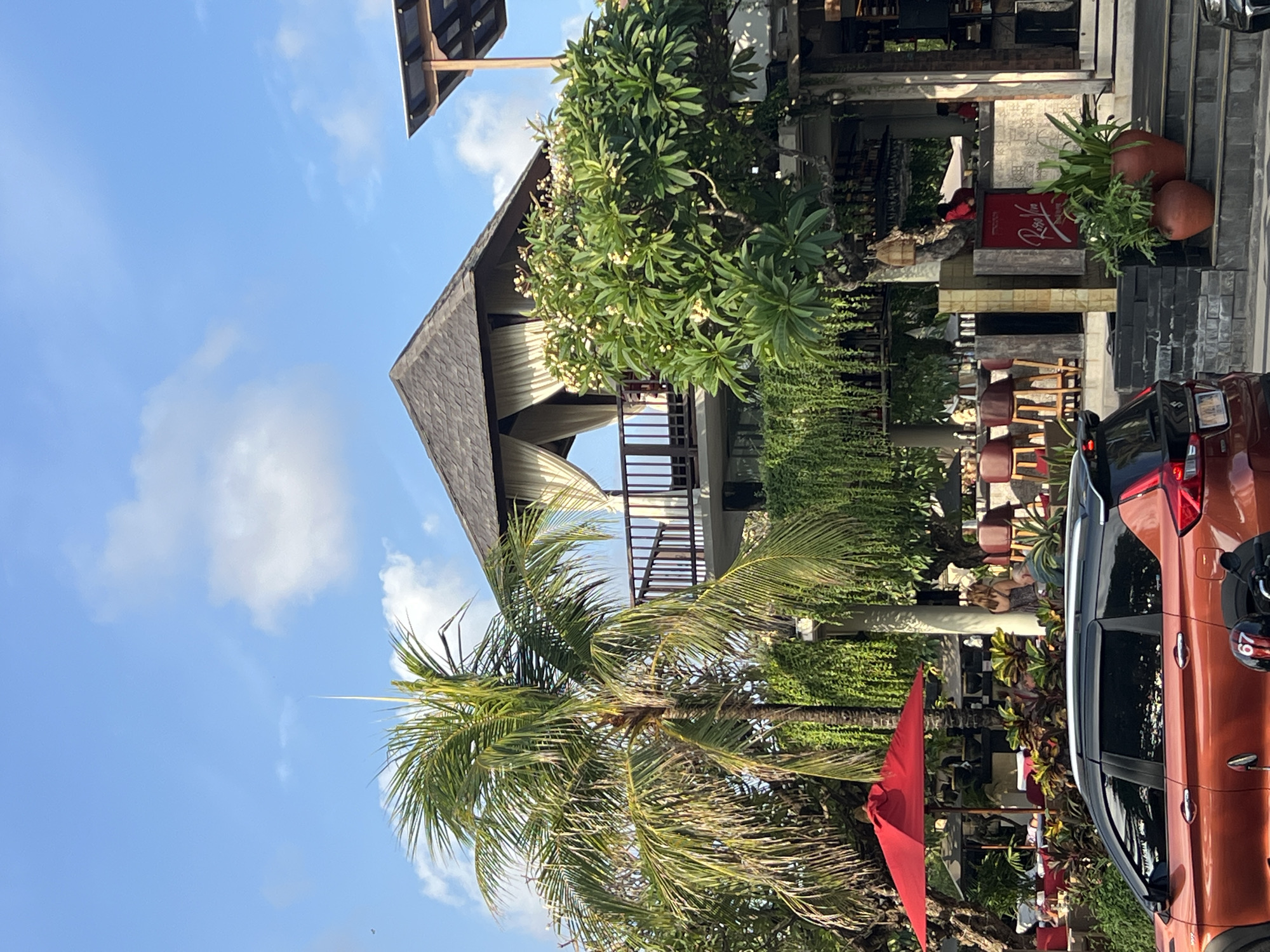 This is a street-level, eye-level, medium shot of a building in Kuta, Indonesia, on a sunny day. A bright orange car is parked in the foreground. Behind the car, there are trees, a red umbrella, and part of a building. The building has multiple levels, with a balcony on the upper level covered by curtains. There are also outdoor seating areas and a bar. Green vines hang from the building, and potted plants are placed near the entrance. The sky is blue with scattered clouds.