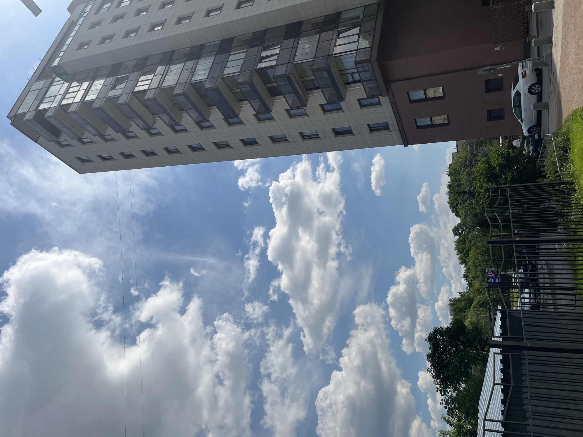 The image captures a view of a modern, multi-story apartment building in Saint Petersburg, Russia, under a bright blue sky dotted with fluffy white clouds. The building, with its light-colored facade and numerous balconies with dark railings, stands tall and dominates the right side of the frame. A lower section of the building, clad in dark red brick, forms its base.
In the foreground, a black metal fence borders an area with lush green grass and vegetation. Beyond the fence, several cars are parked, including a white sedan in the mid-ground. Trees and greenery surround the buildings, suggesting an urban setting with significant parkland or tree-lined streets. The presence of power lines stretching across the sky indicates an established urban infrastructure. The overall scene depicts a sunny day in a residential part of Saint Petersburg.