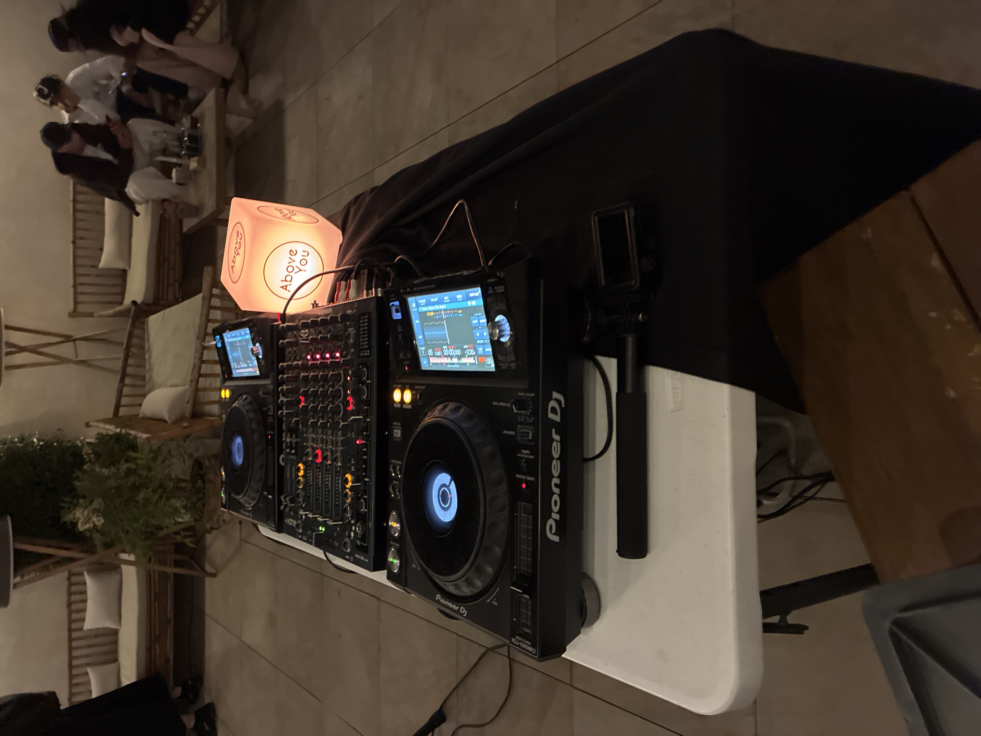 The image shows a DJ setup consisting of two Pioneer DJ CDJ-3000 media players and a DJM-900NXS2 mixer on a white folding table, likely at an event. An illuminated cube with the words "Above You" is positioned next to the setup. A camera on a tripod stands to the right of the media player. The equipment is situated on a tiled surface, with a dark drape partially covering the table. In the background, several seated individuals are visible, suggesting a social gathering. The location is likely an outdoor patio in Barcelona, Spain, identified by the specific "Above You" branding used in several nightlife venues there. The environment suggests an evening event or party.