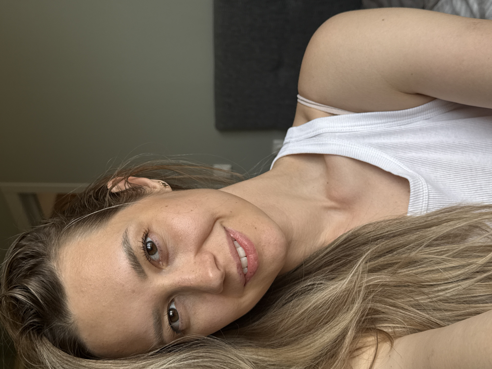 A light-skinned woman with shoulder-length blonde hair is lying down and looking at the camera. She is wearing a white ribbed tank top. Her location is unknown; the image provides no information to identify Chaikovsky, Russia, or any other specific location. The background shows a gray wall and headboard.