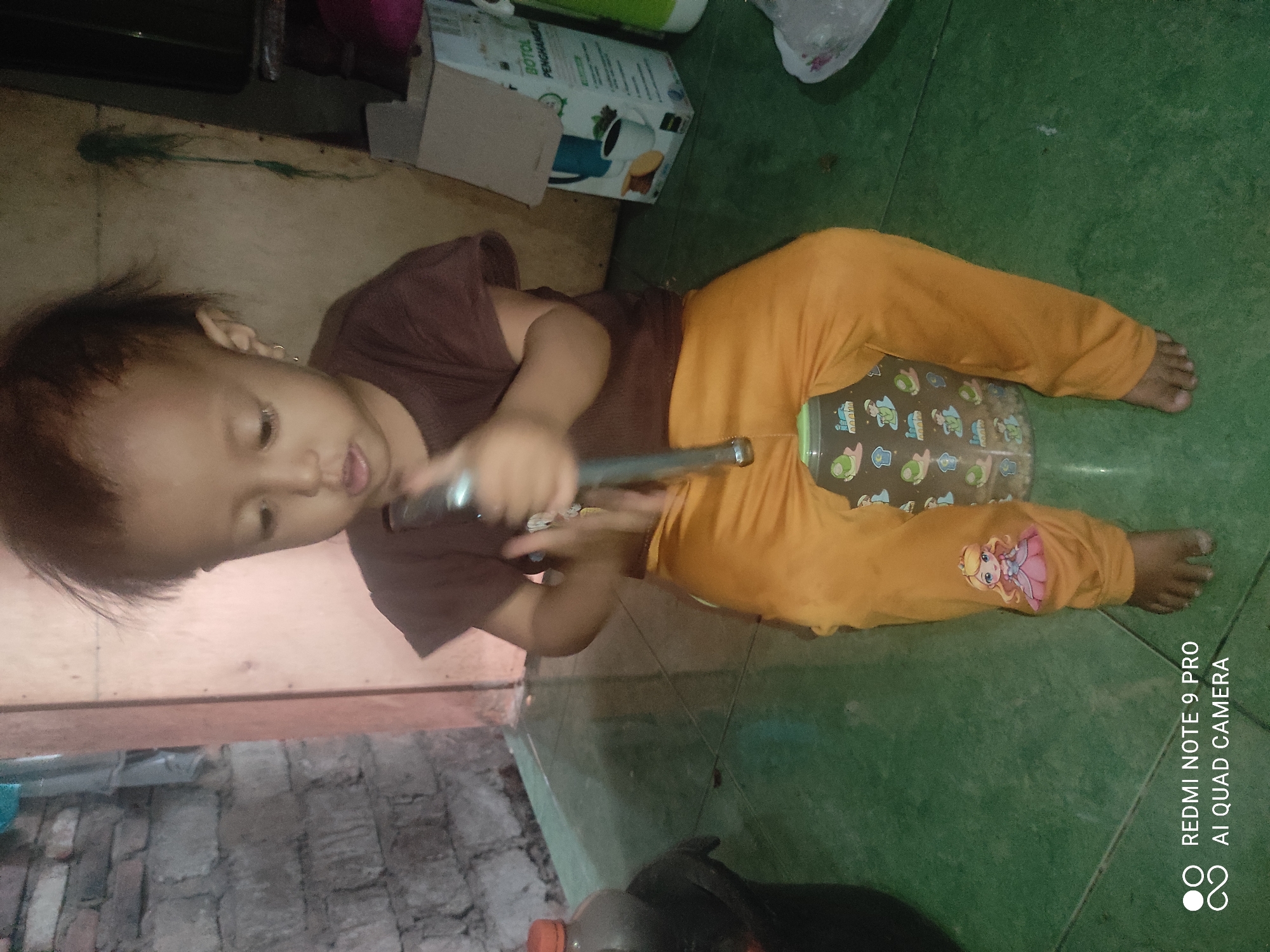 A young child is sitting on a translucent plastic container on a green tiled floor. The child is wearing a brown t-shirt and orange pants with a pink princess decal on the left leg. The child is holding a silver metal tube with both hands. The background consists of a brick wall on the left and a wooden wall on the right. There is a cardboard box with Indonesian text visible on the right side of the frame, and a black plastic container with a red cap on the far left. The lighting is dim. The image contains a watermark in the bottom left corner indicating "REDMI NOTE 9 PRO AI QUAD CAMERA".
