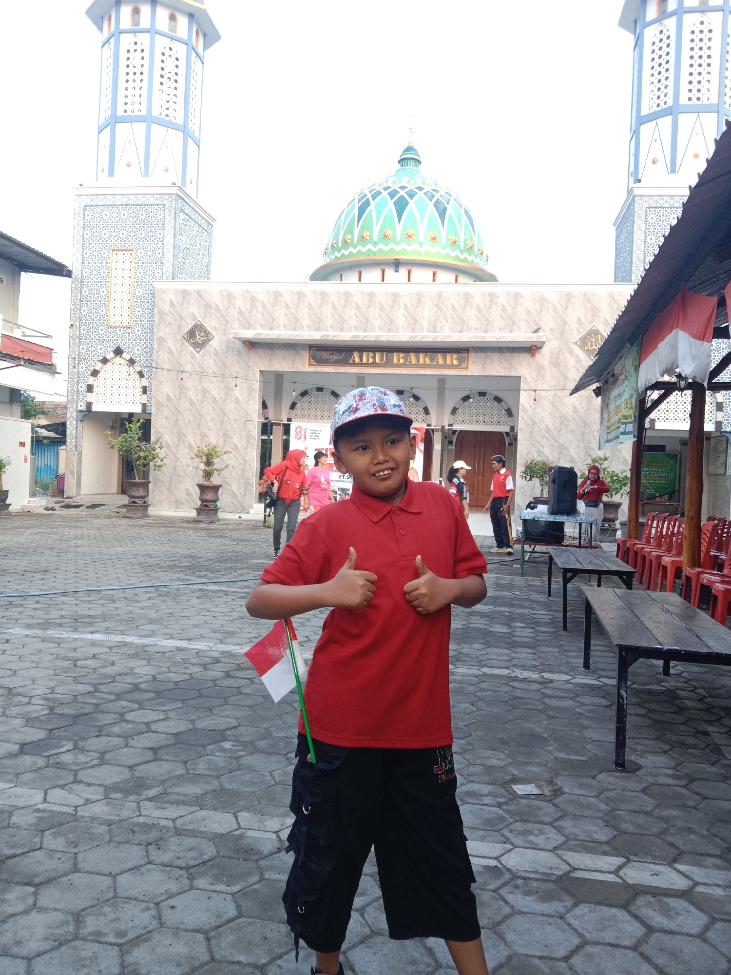 A young boy in a red polo shirt and black cargo shorts, wearing a patterned baseball cap, stands in front of a mosque. He is smiling and giving a double thumbs-up gesture. He is holding a small Indonesian flag. The mosque, named Masjid Abu Bakar, has a prominent green dome and two minarets. The surrounding area is paved with hexagonal stone tiles. In the background, there are other people, tables, chairs, and banners, suggesting an outdoor event or gathering. The overall scene appears to be taking place in Madiun, Indonesia, given the name of the mosque and the presence of the Indonesian flag.