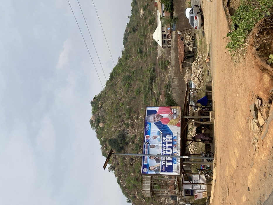 The image depicts an unpaved roadside area in Magongo, Kogi State, Nigeria, against a backdrop of a prominent green and rocky hill under an overcast sky. A large billboard, advertising "Convention 2025: The Way of Truth!" and featuring multiple male figures, including a large portrait of Rev. Dr. Matthew Adelabu, is centrally located on a metal frame structure. Below the billboard, a rustic, open-sided shelter with a corrugated metal roof provides shade; three individuals are seated beneath it, one in light attire, and two others partially obscured in darker clothing. A motorcycle is parked to the left of this shelter.
To the left of the billboard structure, a raised metallic frame supports a water tank. A tall pole with a rectangular solar-powered streetlight fixture is positioned directly in front of the billboard. The foreground consists of reddish-brown dirt, showing ruts and exposed rocks. On the right side of the dirt area, a silver sedan is parked with its trunk slightly ajar. Piles of rocks and debris are scattered across the ground near the shelter. Electrical lines run diagonally across the upper right portion of the image, traversing the hillside. In the middle distance on the right, a distinctive multi-storied building with a white conical roof is visible on the hillside, along with other scattered, less defined structures. Sparse green vegetation covers the hill and parts of the immediate foreground.