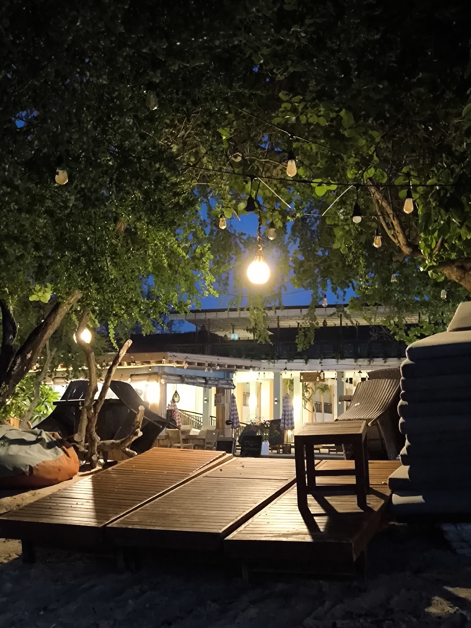 The image depicts an outdoor seating area on a beach in an unknown location in Indonesia at night. The area is illuminated by string lights hanging from trees. Wooden lounge chairs are arranged on the sandy ground. A small table is placed next to a stack of towels. In the background, a building with outdoor seating can be seen. The overall scene creates a relaxed and inviting atmosphere.