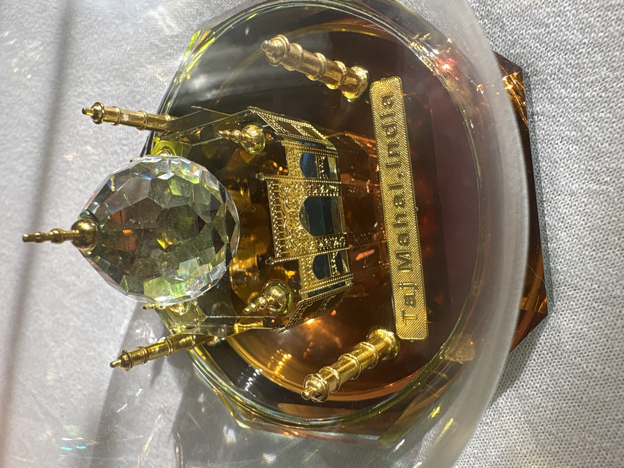 A souvenir of the Taj Mahal, India, enclosed in a clear plastic dome, sits atop a light gray textured surface. The souvenir is predominantly gold, with a small model of the Taj Mahal structure next to a multifaceted crystal dome. A gold-colored plate inscribed with "Taj Mahal, India" in vertical orientation is also inside the dome. The souvenir is made of metallic and plastic elements, with reflections inside the dome creating highlights and patterns. The base of the souvenir is a dark amber or reddish-brown color. The city of Tri Protoka, Russia, is not visible in this image.