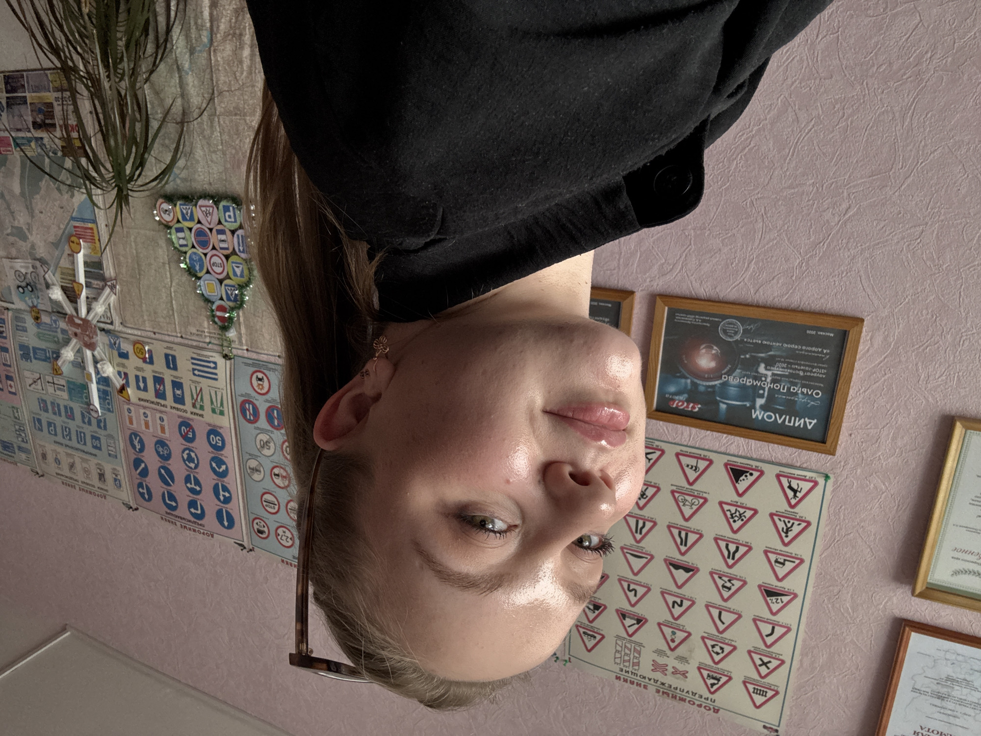 The image depicts a young woman with blonde hair and fair skin, wearing a black jacket. She is looking towards the camera with a slight smile. Her face is illuminated, highlighting her features. In the background, a wall is covered with numerous road signs, displayed in a triangular arrangement, resembling a Christmas tree. Additional road signs are visible on the wall, along with framed documents and a potted plant. The room appears to be decorated with a theme of traffic safety and driving education. Based on the visible text on the framed documents and the common educational materials found in driving schools, it is highly probable that this scene is located in the city of Chaikovsky, Russia, likely within a driving school or an educational institution.
