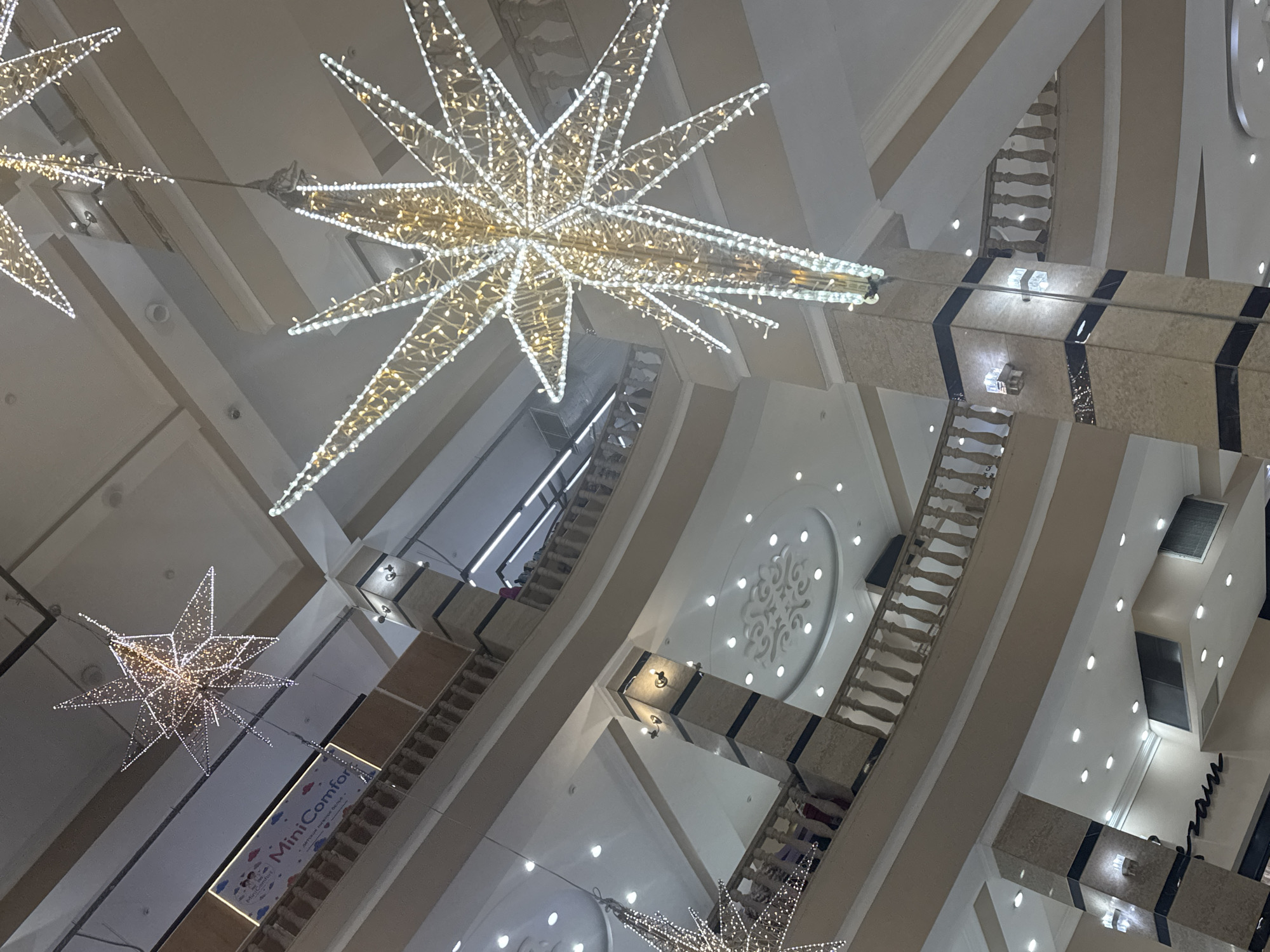 The image shows the interior of a multi-level shopping mall decorated with illuminated star-shaped ornaments. The architecture features beige walls, columns with marble and black accents, and ornate railings on the balconies of each level. Multiple ceiling lights are visible, along with what appears to be air conditioning units. On the left, a sign for "MiniComfort" is partially visible. People can be seen on the upper levels, near the railings. The overall impression is of a well-lit, contemporary indoor shopping space, likely decorated for a festive occasion. The location is identified as Bishkek City, Kyrgyzstan.
