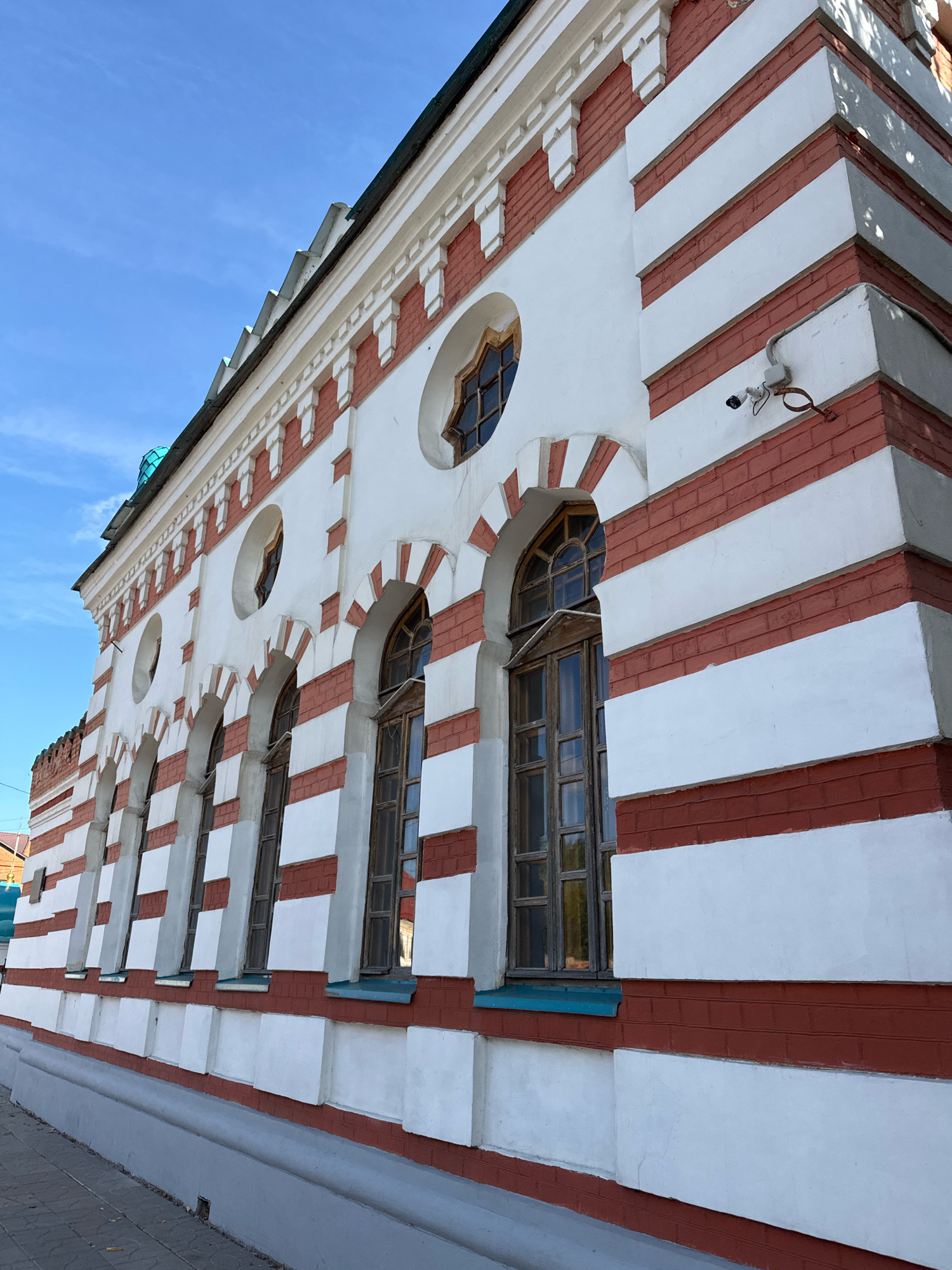 The image shows a building with red and white striped walls, located in the city of Tver, Russia. The building has a series of arched windows with wooden frames and a decorative cornice. A security camera is mounted on the exterior wall. The sky is clear and blue. The building appears to be in good condition, with no visible signs of damage or neglect. The overall scene is one of architectural interest and historical significance.
