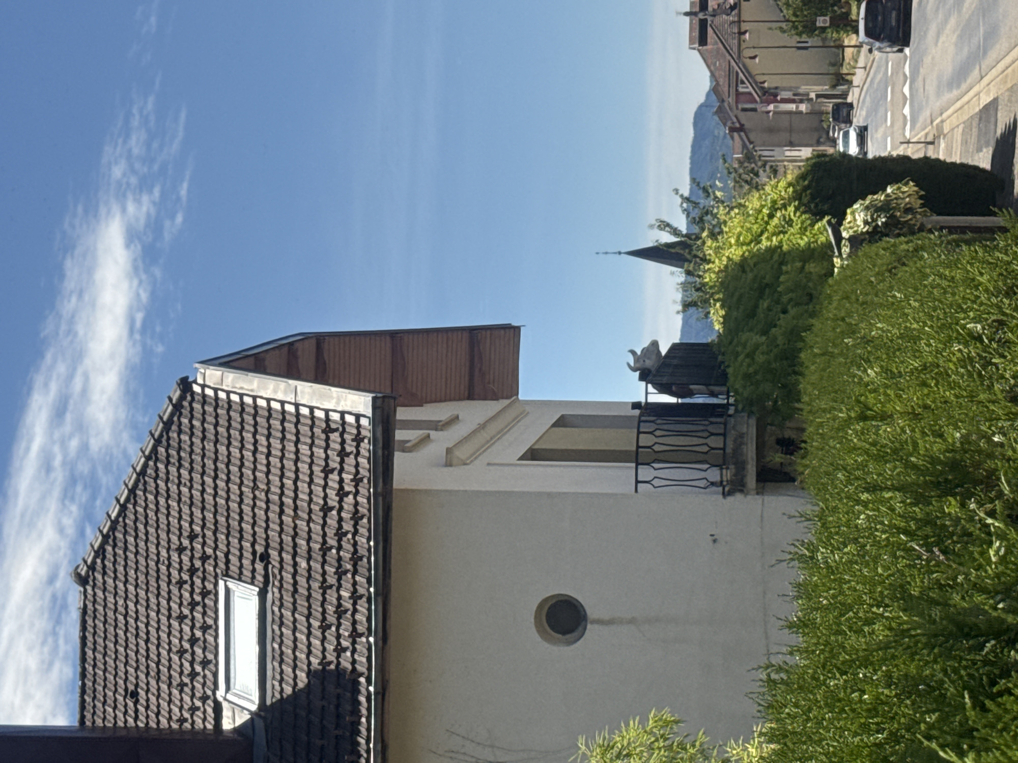 A street scene in Coteaux du Lizon, France, under a clear blue sky with scattered white clouds. The foreground is dominated by a dense, green hedge. Behind the hedge, a white building with a dark tiled roof and a dormer window is partially visible. A round window is set into the building's facade, and a balcony with a black metal railing overlooks the street. A statue of a cow's head with horns is positioned on top of a dark wooden structure on the balcony. Across the street, a paved road with a pedestrian crossing leads through a residential area lined with buildings. Several cars are visible on the road, and a church spire rises in the distance. The overall scene suggests a sunny day in a French town.