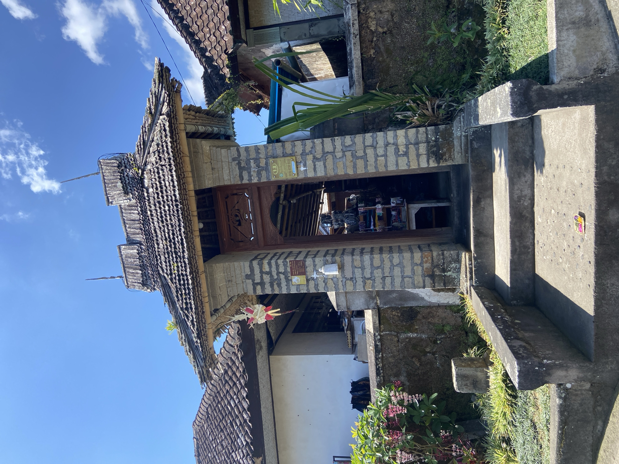 The image captures an entrance to a small shop in Bangli, Indonesia, under a bright blue sky with scattered clouds. The entrance is framed by a stone archway supporting a weathered, tiled roof with wooden trim. A traditional Balinese offering, a "canang sari," made of palm leaves and flowers, hangs from the roof's overhang. Stone steps lead up to the entrance, flanked by low stone walls and lush green vegetation, including flowering plants with pink blossoms and tall green stalks. The shop itself appears to be open, with shelves stocked with various items visible through the doorway. The buildings surrounding the entrance have red-tiled roofs and white walls, typical of Indonesian architecture. The overall scene conveys a sense of local commerce and traditional culture in a tropical setting.