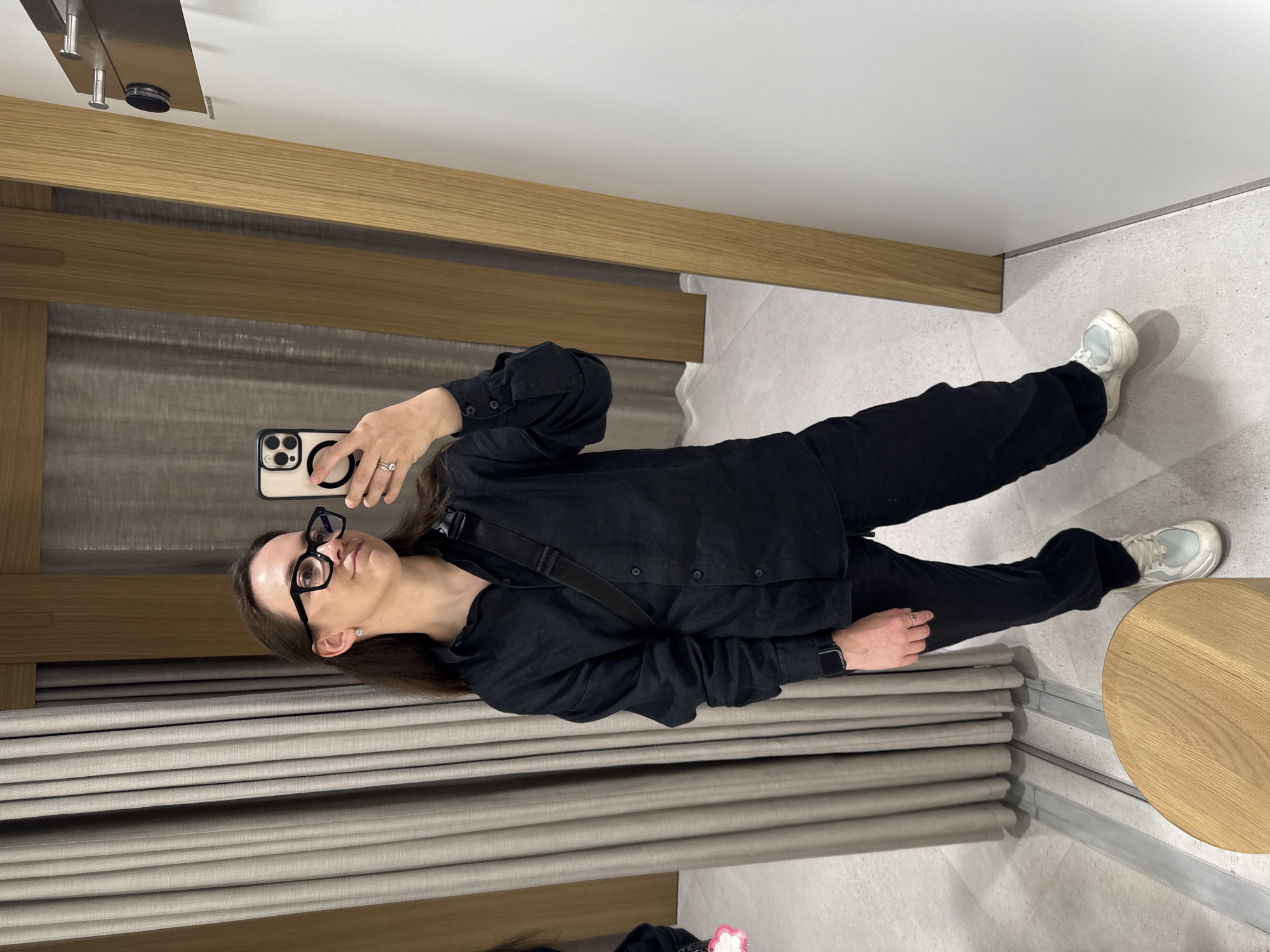 A woman with long dark hair and wearing black-framed glasses, a black shirt, black pants, and white sneakers is taking a selfie in what appears to be a fitting room. She is holding a smartphone with a visible camera setup. The room has a wood trim along the upper wall and a gray curtain as the backdrop. A small round table is visible on the right. The image does not have any information about the location in Nizhny Novgorod, Russia.