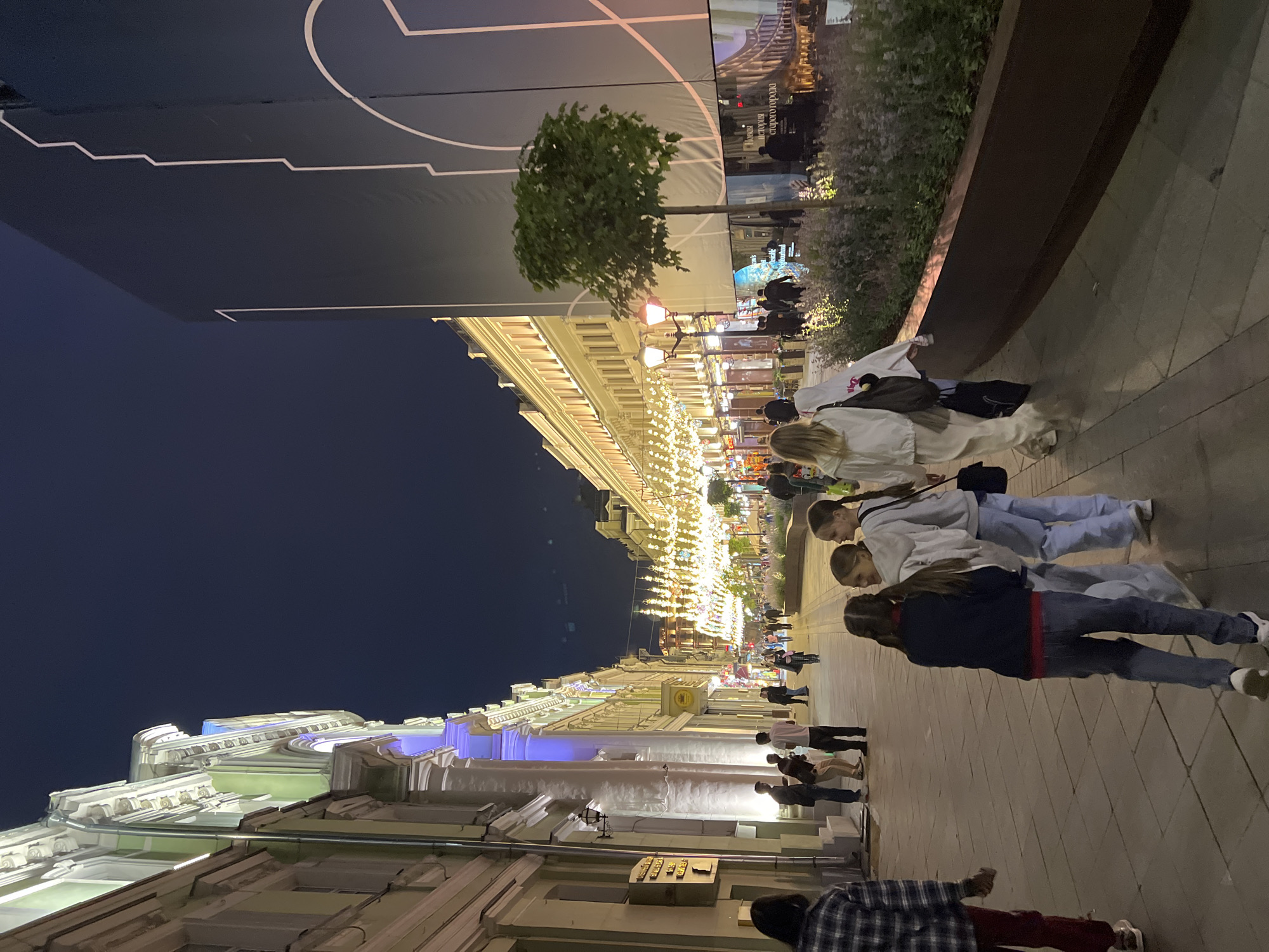 The image depicts a street scene in Moscow, Russia, at night. The street is illuminated by decorative lights strung across the pathway and the buildings. On the left side of the street, there are ornate buildings with illuminated architectural details. On the right side, a large dark banner with white geometric lines is displayed, possibly for an advertisement or event. There are several people walking along the street. In the foreground, a group of four young females, appearing to be teenagers, are walking away from the camera, two of them are in conversation. Further down the street, other pedestrians are visible, and shops are lining the buildings. The atmosphere appears to be lively, with the lights creating a festive ambiance. The location is likely a well-known pedestrian street in Moscow, possibly Arbat Street or a similar area, given the architectural style and decorative lighting.