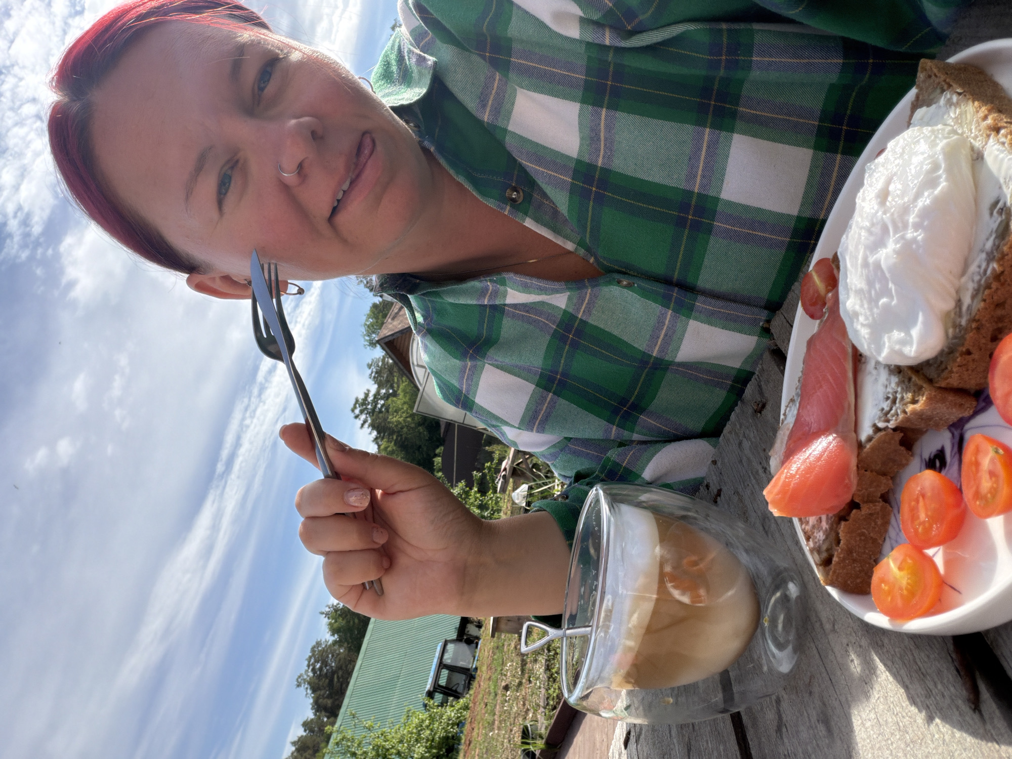 The image shows a woman holding a fork, with a plate of food and a glass of drink on a wooden surface. The food appears to be salmon, tomatoes, and a poached egg on bread. The drink in the glass is a light-colored liquid with visible layers. The woman is wearing a green and white plaid shirt and has red hair. In the background, there is a green roof, trees, and a sky with clouds. The location is in the city of Chaikovsky, Russia.