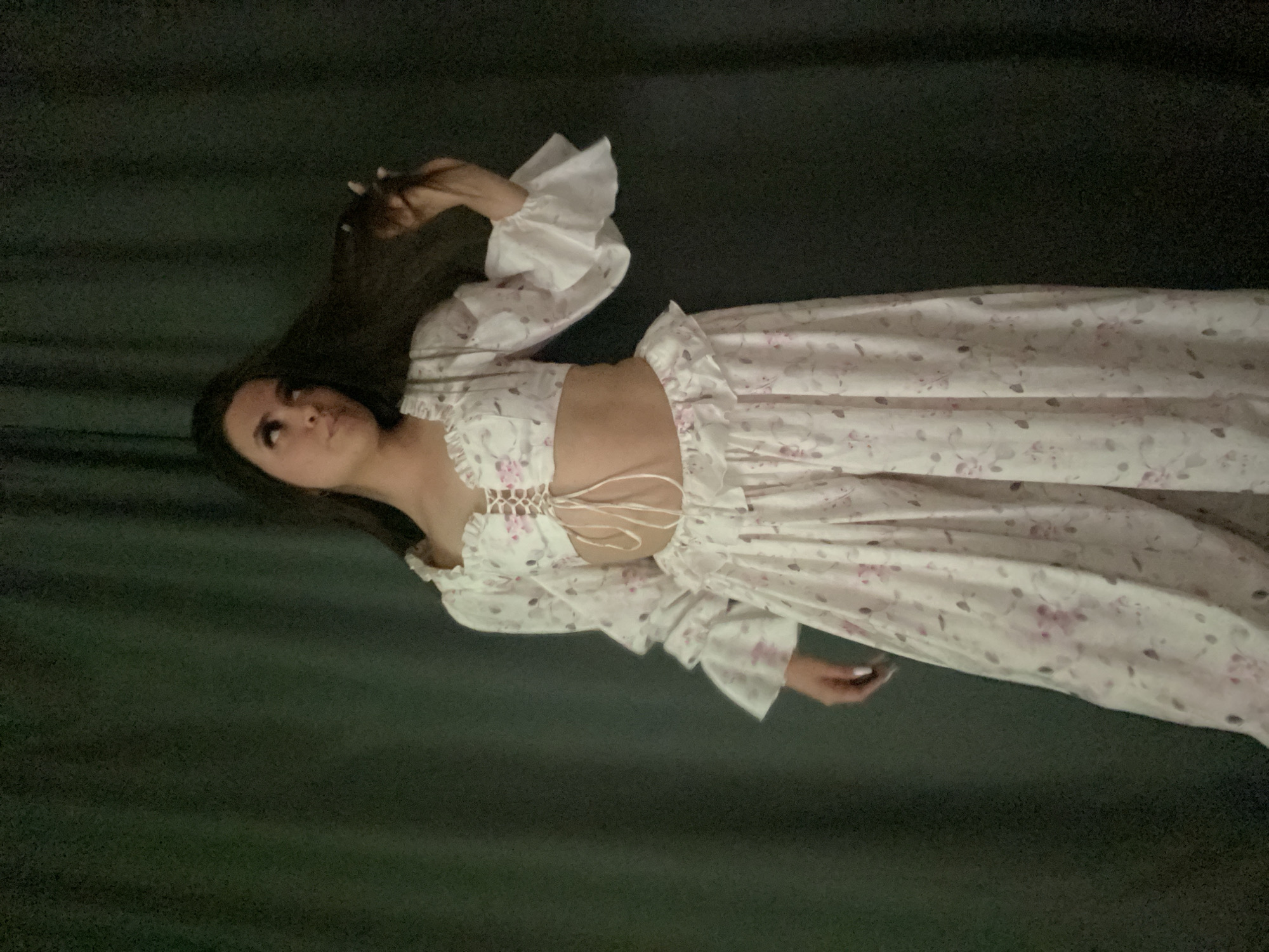 A woman wearing a floral dress with a corset-style top and ruffled sleeves is lying on a dark, solid-colored surface, likely a bed or couch. Her dark hair is spread out on the surface. The location is unknown.